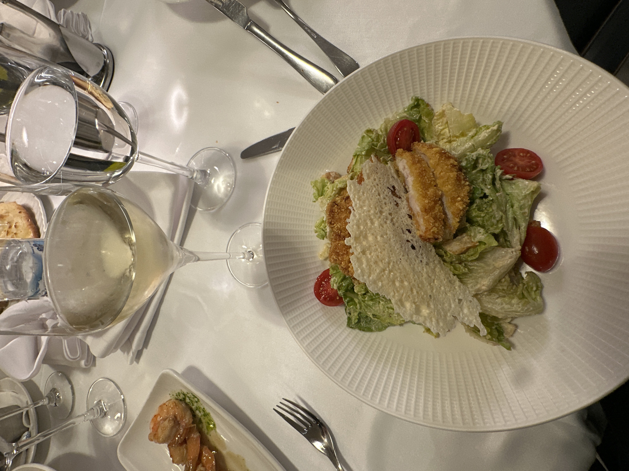 A white table setting features a plate of Caesar salad with fried chicken, croutons, lettuce, and sliced tomatoes. A glass of white wine and a glass of water are present, along with silverware, including knives and a fork. A small white plate with what appears to be shrimp and a green sauce is also visible. The setting suggests a meal in a restaurant, but the location and city are unknown.