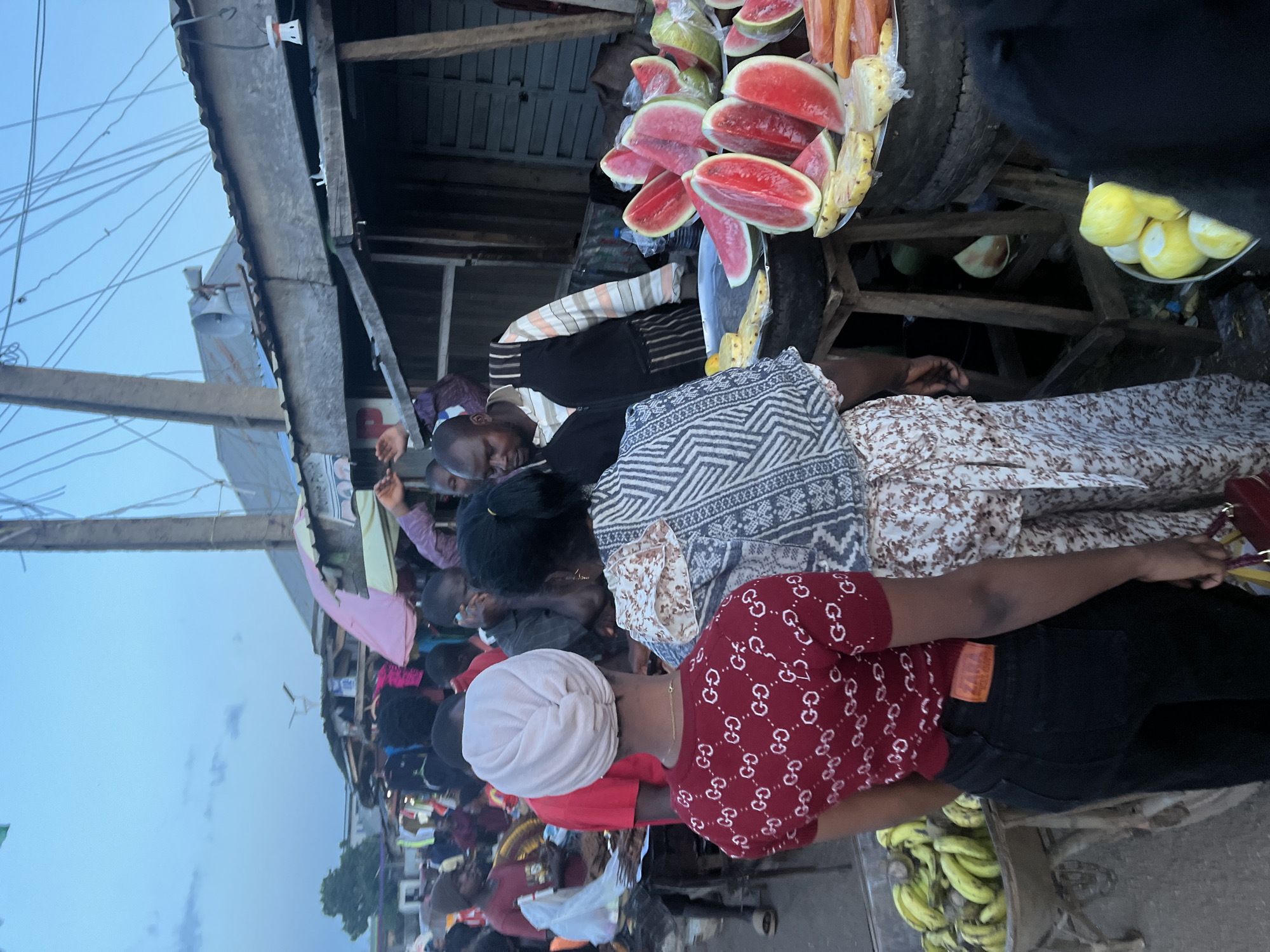 A bustling marketplace scene unfolds in Gura Top, Nigeria, under an overcast sky. The image captures a moment of commerce, with several individuals engaged in transactions. In the foreground, a woman wearing a red patterned shirt and black pants stands facing away from the camera, her back to the viewer. To her right, another woman in a patterned dress is partially visible.
Behind them, a man in a striped shirt and vest stands by a stall laden with cut watermelons and pineapples. He appears to be attending to customers, as implied by the presence of other individuals browsing the produce. The marketplace is characterized by makeshift stalls constructed from wood and corrugated metal, suggesting a dynamic and active commercial hub.
Various items are displayed for sale, including fruits like bananas, watermelons, and pineapples, as well as what appear to be clothing items in the background. The overall atmosphere is one of vibrant activity and daily life in a Nigerian market.