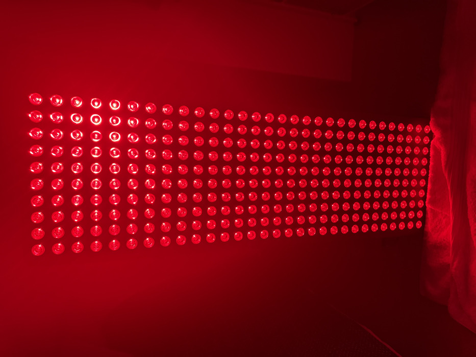 The image displays a vertically oriented rectangular panel densely populated with uniformly arranged, illuminated red circular lights, set against a dark red background. The panel consists of approximately ten columns and over forty rows of individual light sources, casting an intense red glow throughout the scene. Below the panel, a surface covered with a folded, soft, red-illuminated textile material is visible. There are no individuals, subjects, or actions present. No visual cues within the image indicate its location as the city of London, United Kingdom, or any other specific geographical place.