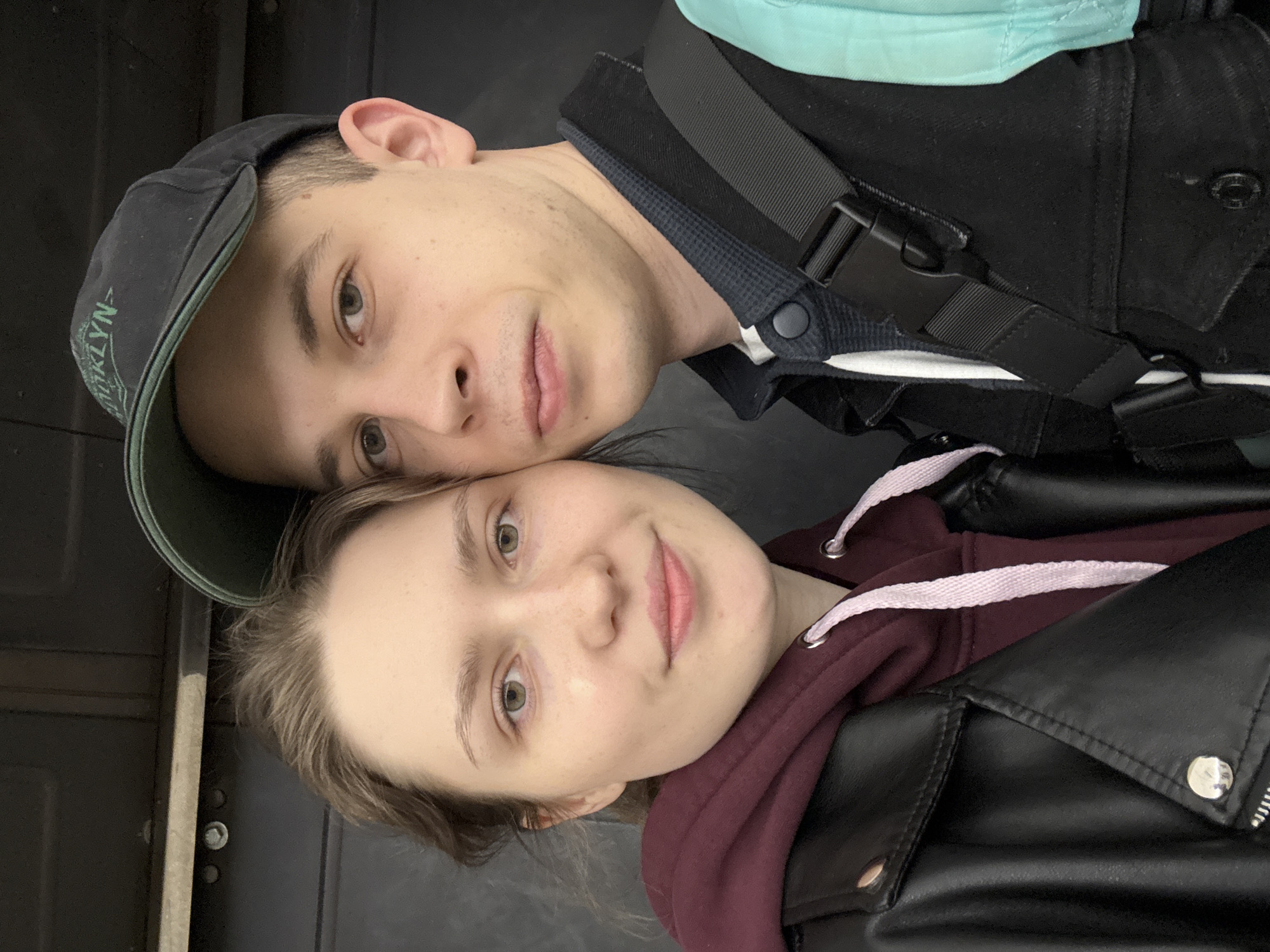 A couple is posing for a selfie in what appears to be an indoor setting. The man is wearing a dark baseball cap with the word "BROOKLYN" visible on the side, a dark shirt with a buttoned collar, and a black bag strap across his chest. The woman is wearing a maroon hoodie under a black leather jacket. The background is nondescript, with dark grey or black surfaces visible. Based on the style of the photograph and the apparent casual setting, it is not possible to discern a specific location in Saint Petersburg, Russia without additional context.