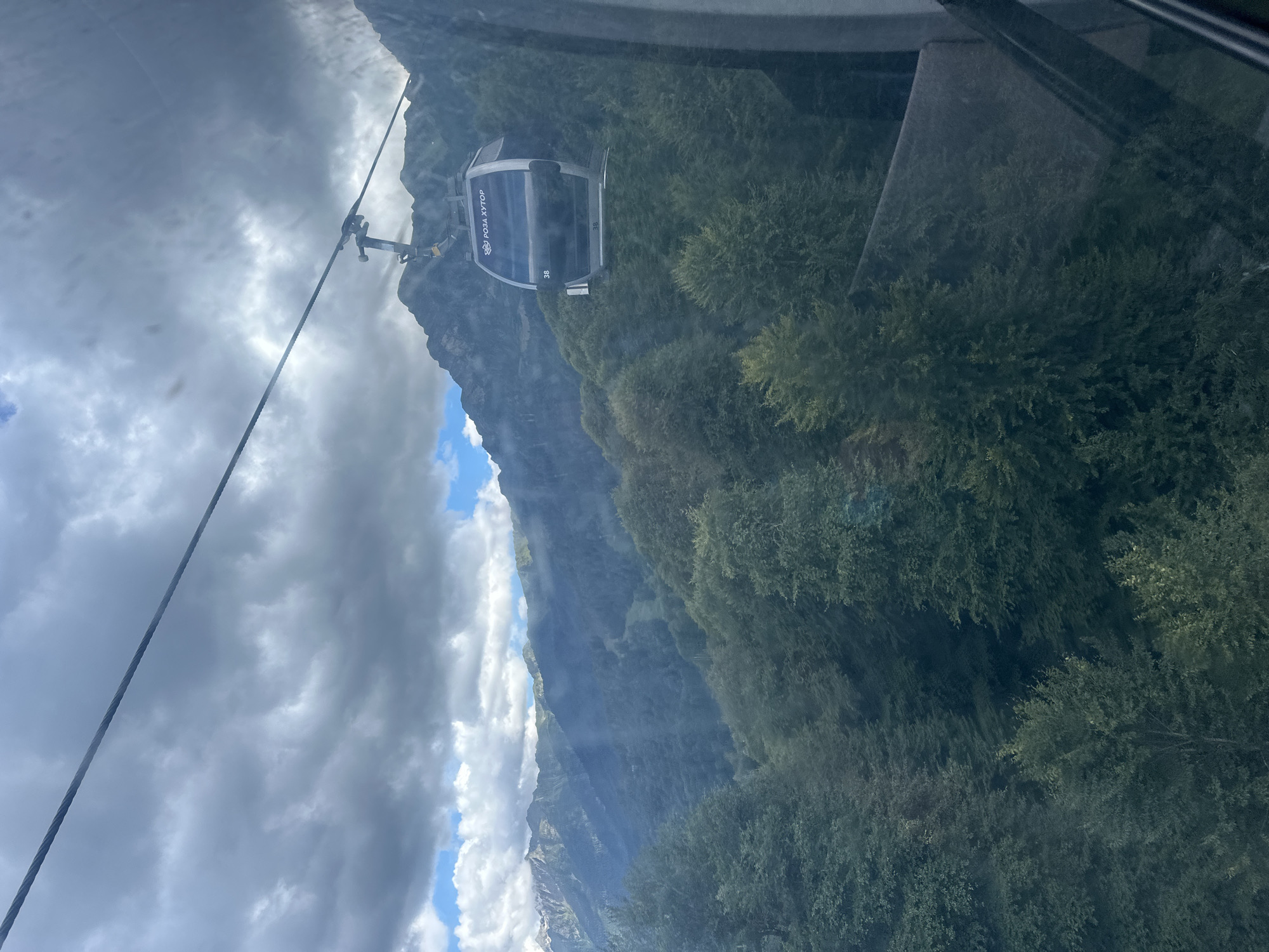 A cable car is seen traveling through a mountainous landscape. The cable car is dark gray and has the inscription "РОЗА ХУТОР" in white lettering on its front. The number "38" is also visible on the cable car. The cable car is suspended from a thick steel cable that stretches diagonally across the frame. Below the cable car, a dense forest of green trees covers the steep slopes of the mountains. The mountains themselves are rugged and imposing, with some of their peaks obscured by dramatic, cloudy skies. Patches of blue sky are visible through the clouds. The scene appears to be captured from within another cable car, as a window frame is visible on the right side of the image. The location is identified as Estosadok, Russia, which is known for its ski resorts and mountain views.