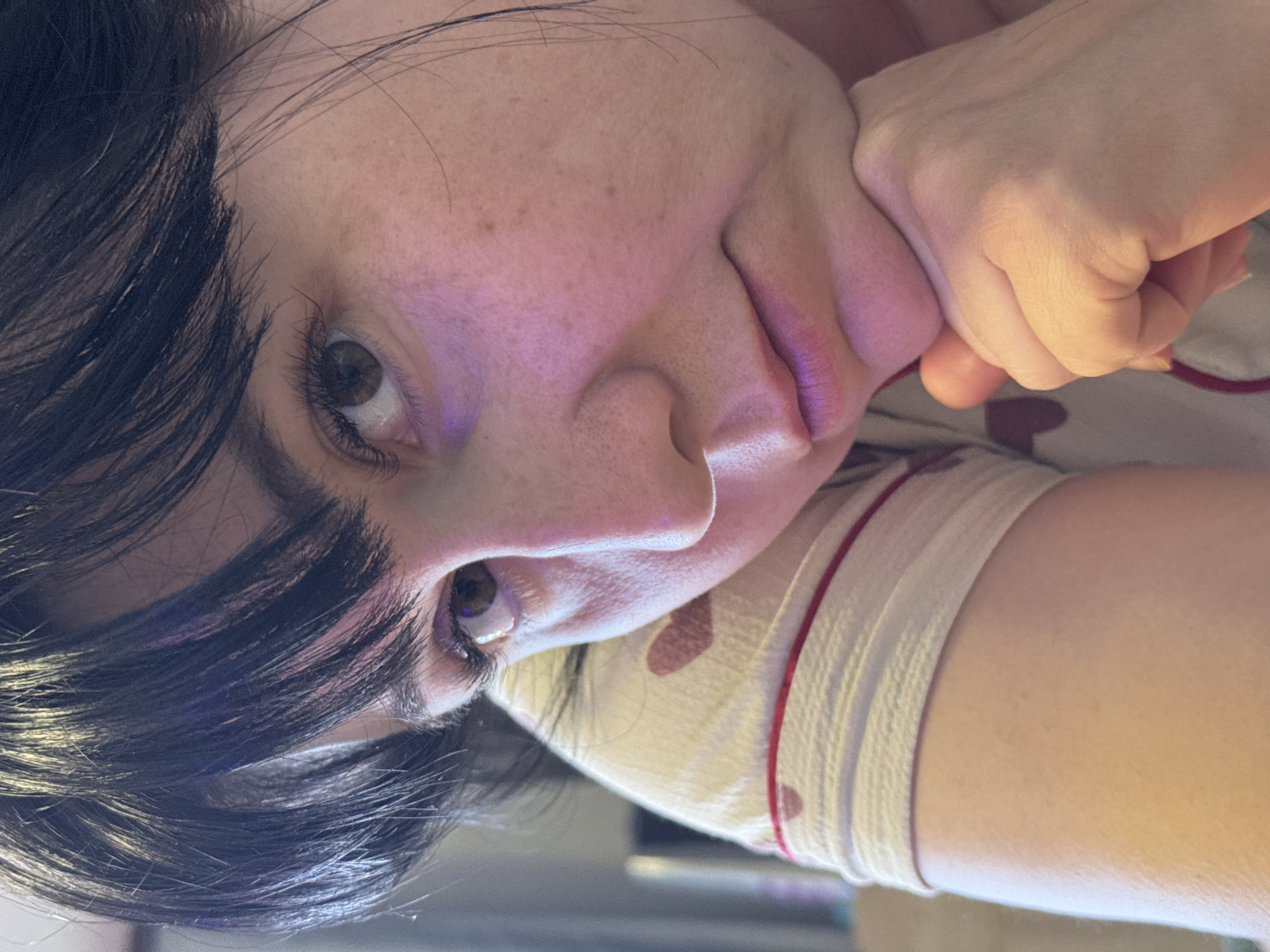 A close-up, portrait-oriented image shows a woman with dark hair, brown eyes, and fair skin. She is resting her face on her hand, her features prominent. Her hair is layered with some strands falling across her forehead. She is wearing a light-colored top with a pattern of red hearts and a red trim around the collar. The lighting appears to be artificial, with a slightly warm hue on her skin. The background is indistinct but seems to suggest an indoor environment. No location information about Zav'yalovo, Russia, is present.