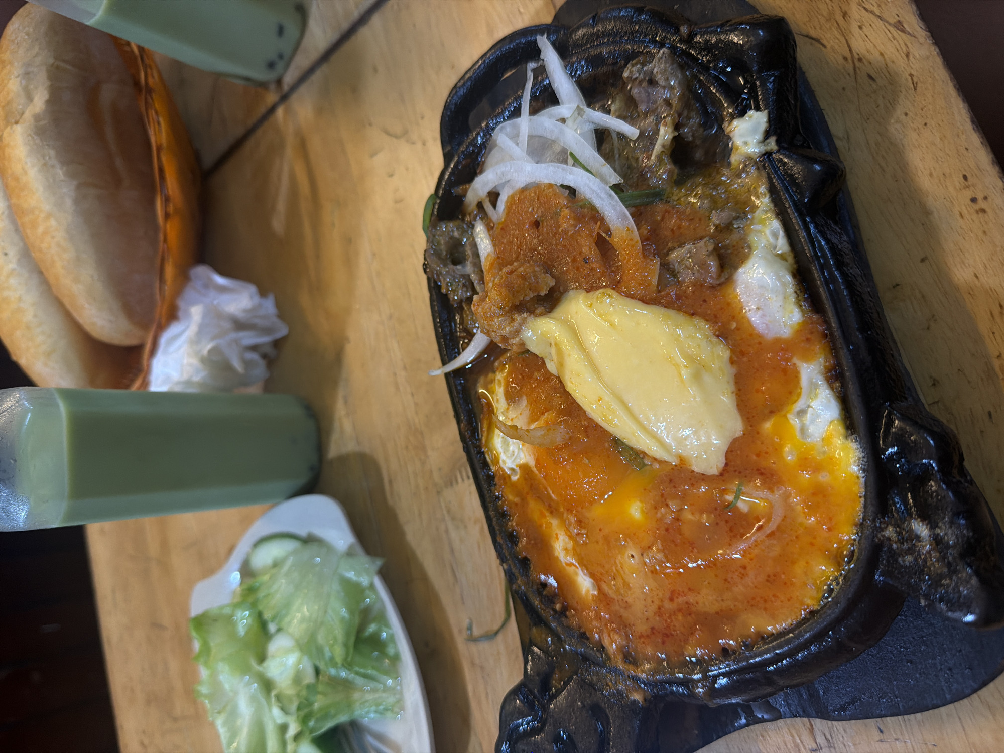 This image captures a meal served on a light-colored wooden table within an establishment situated in Nha Trang Ward, Vietnam. In the foreground, a black, oval-shaped cast iron sizzling plate presents a dish featuring two fried eggs with prominent orange yolks, cubed meat (identifiable as beef), a reddish-orange savory sauce, and a centralized dollop of yellow butter. Slices of white onion and green garnishes are integrated into the dish. To the immediate left, a white ceramic plate holds a side of fresh green lettuce and sliced cucumber. In the upper background, a woven basket contains two light brown, elongated bread rolls. Two clear plastic bottles are visible, one prominently on the left and another partially cropped on the right, each containing a pale green beverage with dark sediment. The wooden table displays visible grain and minor surface imperfections. No individuals are present in the scene.