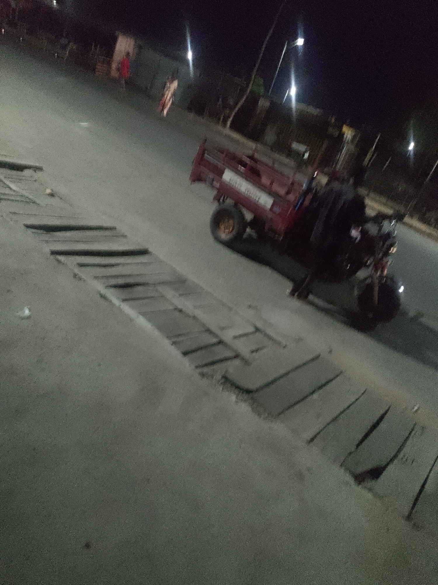 The image depicts a nighttime scene on a paved road in Jalingo, Nigeria. In the foreground, a three-wheeled vehicle, possibly an auto-rickshaw, is parked on the right side of the road. It is dark red and appears to have a cargo bed. A person, wearing dark clothing, is seated on the vehicle. The road surface shows some wear, with wooden planks or slabs laid across a drain or ditch on the left side of the image.
Further in the background, to the left, two figures are visible walking. One is wearing red, and the other is in a light-colored garment with dark stripes. The setting appears to be a street with some buildings and streetlights visible in the distance, contributing to the nighttime illumination. The overall atmosphere is that of a typical urban street at night. No specific event or activity is clearly discernible beyond the presence of people and the parked vehicle. The lighting suggests it is likely after dark, with artificial light sources creating some glare. The image quality is somewhat grainy, characteristic of low-light photography.