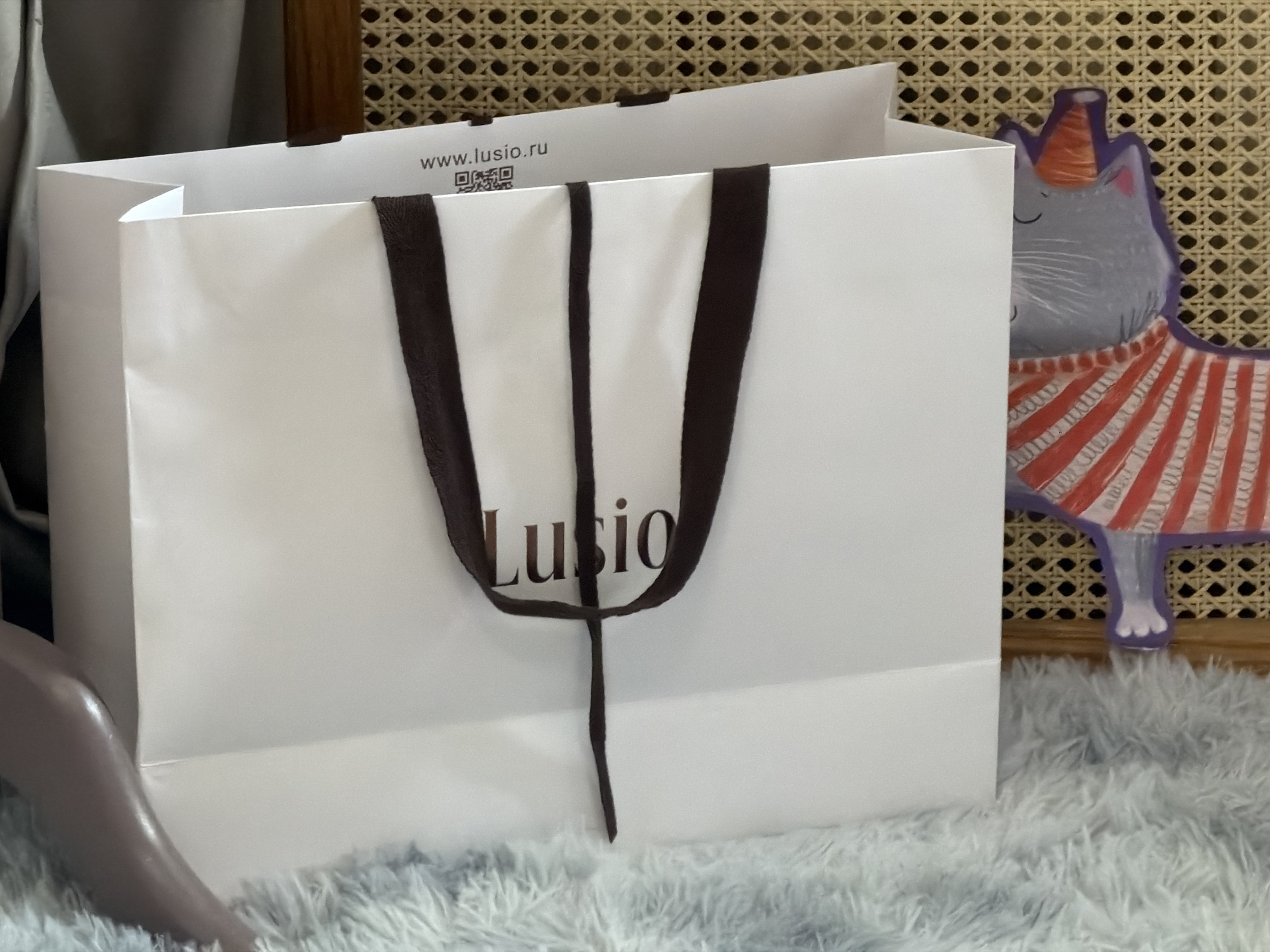 A white shopping bag from the brand "Lusio" is positioned in the center of the frame, resting on a fluffy, light blue surface. The bag has dark brown handles and the brand's website printed on the top edge. A light grey object is visible to the left of the bag. Behind the bag is a wicker surface and a cutout figure of a grey cat with a birthday hat and a red and white striped shirt. The location cannot be determined from the image, however the URL on the bag indicates that the bag is from a company in Russia.