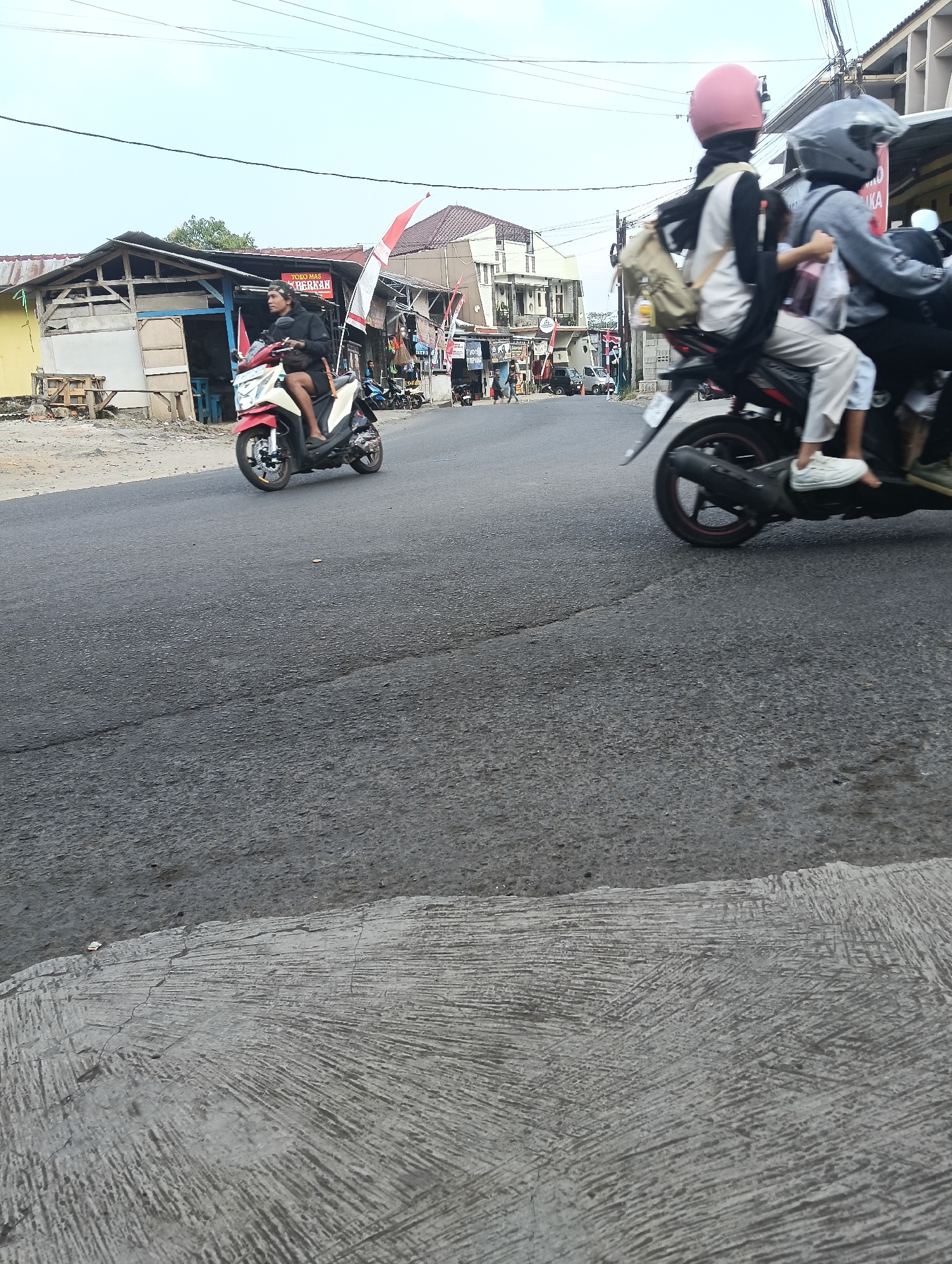 The image depicts a street scene in Puspahiang, Indonesia, with a focus on two motorcycles. The motorcycle in the foreground, on the right, carries two individuals. The person in the rear is wearing a pink helmet and a black hijab, with a tan backpack slung over their shoulders. The person in front is wearing a gray helmet and a gray long-sleeved shirt with white pants. Both are seated on the motorcycle.
To the left, another motorcycle, primarily red and white, is being ridden by a single individual wearing a dark jacket and a headband. This motorcycle is positioned in front of a weathered building with an open front, displaying a sign that reads "TOKO MAS" (Gold Shop) and "BERKAH". In the background, more buildings and a clear sky are visible. The street itself is paved and shows signs of wear. The overall impression is a typical street in a Indonesian town, with everyday activity.