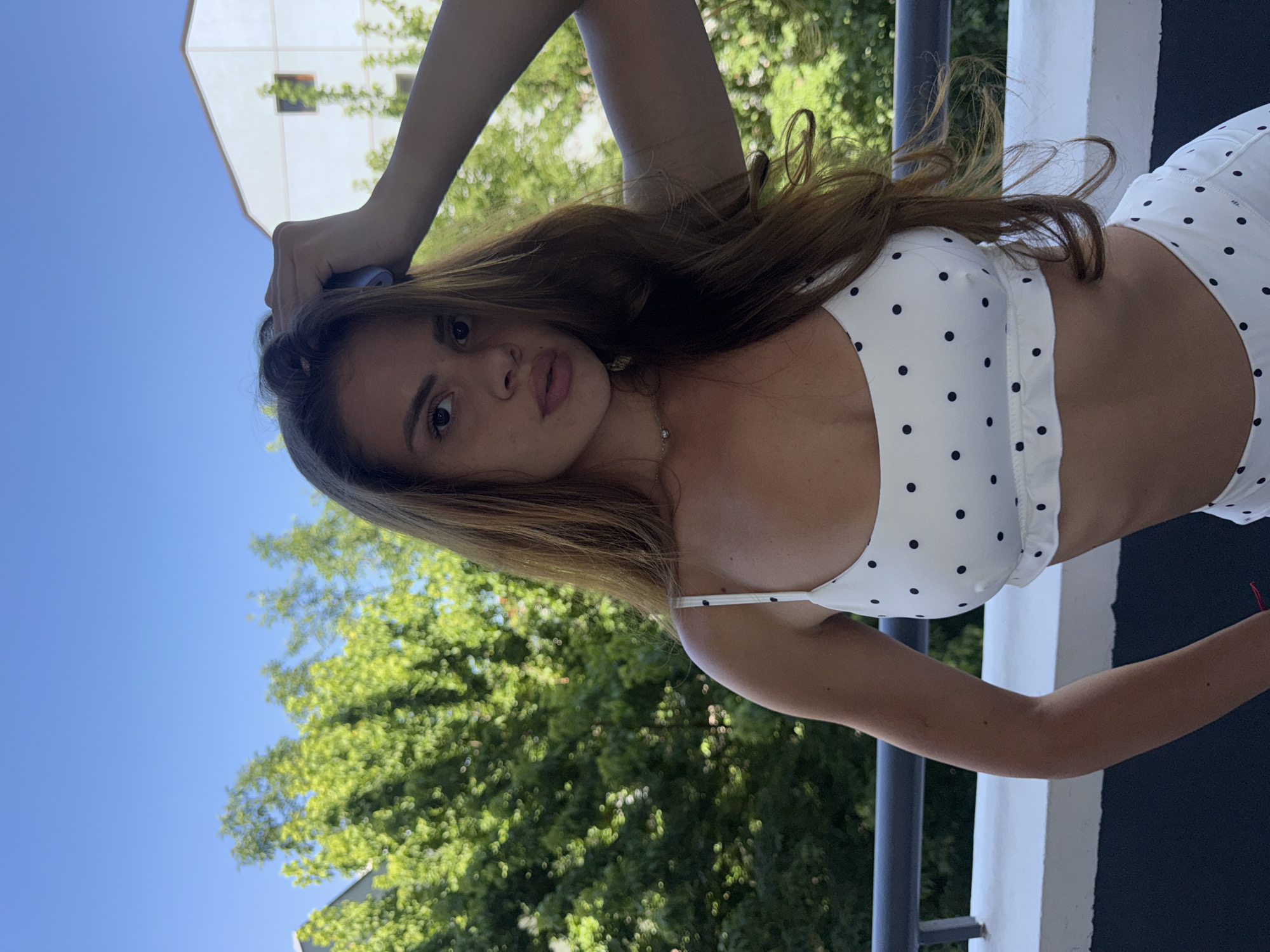 A young woman with long, auburn hair is captured in a low-angle shot, outdoors. She is wearing a white polka-dot bikini top. Her right arm is raised and bent at the elbow, with her hand resting on the back of her head. Her left arm is extended downwards, with her hand resting on a black railing. The woman's face is turned towards the camera, with a neutral expression.
Behind her, lush green trees are visible, interspersed with bright sunlight, suggesting a warm, sunny day. A portion of a white building with a few windows can be seen in the upper left corner of the frame. The sky above is a clear, vibrant blue. The scene appears to be a balcony or terrace, given the railing and the perspective looking upwards. There is no visible text in the image. The overall impression is casual and relaxed, likely taken during daylight hours in a location with abundant greenery.