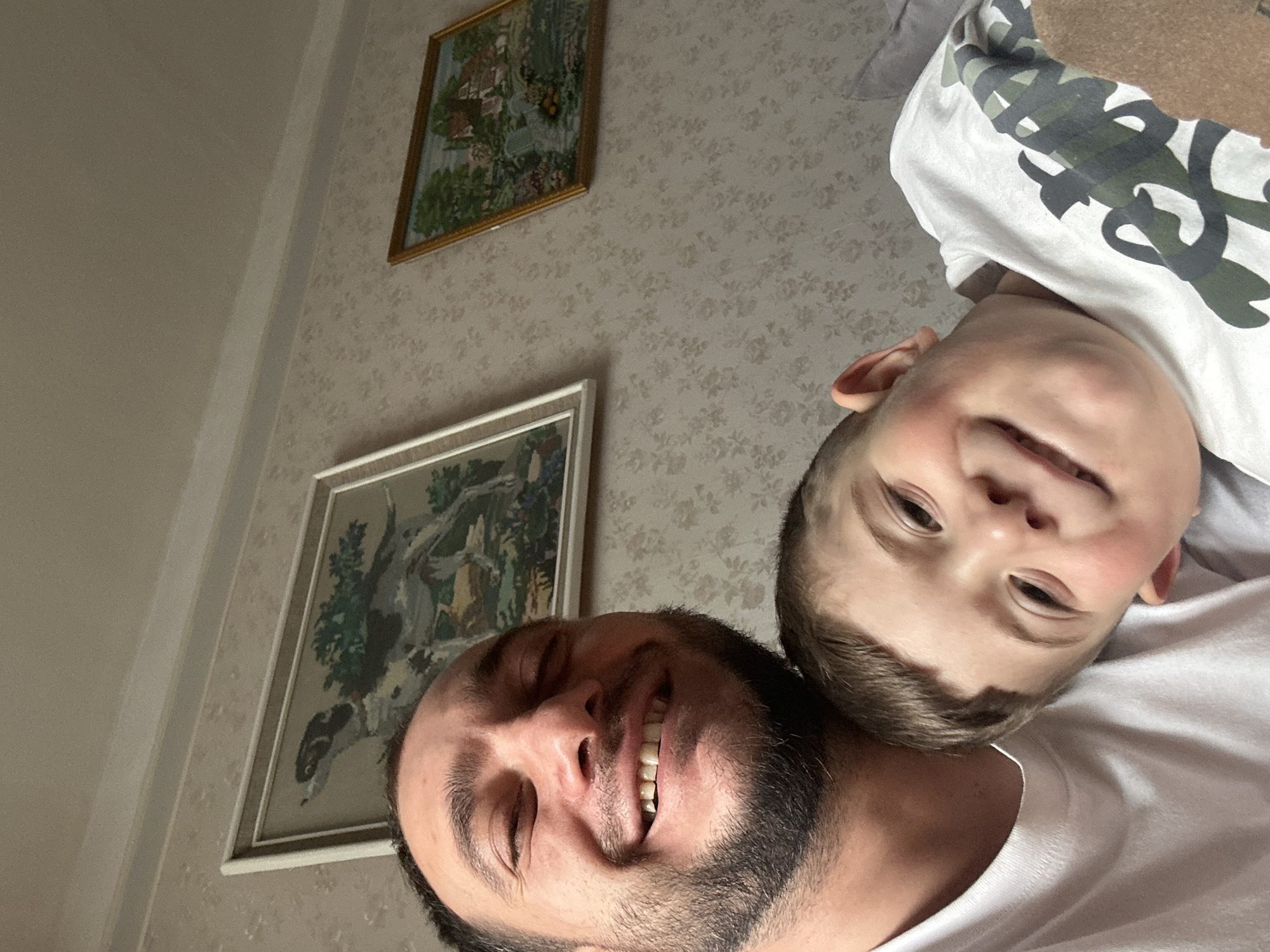 A man and a boy are pictured in a selfie-style shot. The man has dark hair and a beard, and is smiling. The boy has light brown hair and is also smiling. The boy is wearing a white t-shirt with a camo print, and the man is wearing a plain white t-shirt. Two framed pictures are visible on the wall behind them, which has patterned wallpaper. The photo was not taken in the city of Yerevan, Armenia.