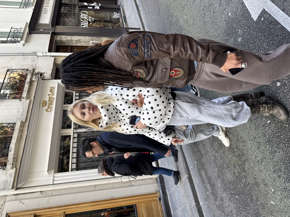 The image depicts a street scene in Paris, France, identifiable by the presence of the shop "Crazy Lou," located at 12 Rue de Turbigo, 75001 Paris. Four individuals are walking on a paved street beside multi-story stone buildings with iron balconies and potted plants. In the immediate foreground, a fair-skinned woman with blonde hair, wearing a white polka-dotted puffer jacket, grey sweatpants, and dark buckled boots, walks with a lit cigarette in her right hand and a smartphone in her left. She faces slightly towards a person walking next to her, on her right. This individual has dark dreadlocks, wears a brown leather bomber jacket adorned with multiple patches (including "SIGNAL CORPS U.S.N. STRONG TO NONE"), brown sweatpants, and holds a smartphone in their left hand. Behind these two, a fair-skinned man with dark, curly hair and eyeglasses wears a dark jacket, light grey pants, and white sneakers. Further back, another person in a dark jacket and blue jeans is partially visible. The environment includes additional shopfronts like "KIFF & MAR" and "SALOTTO pop up." The scene is illuminated by diffused daylight.