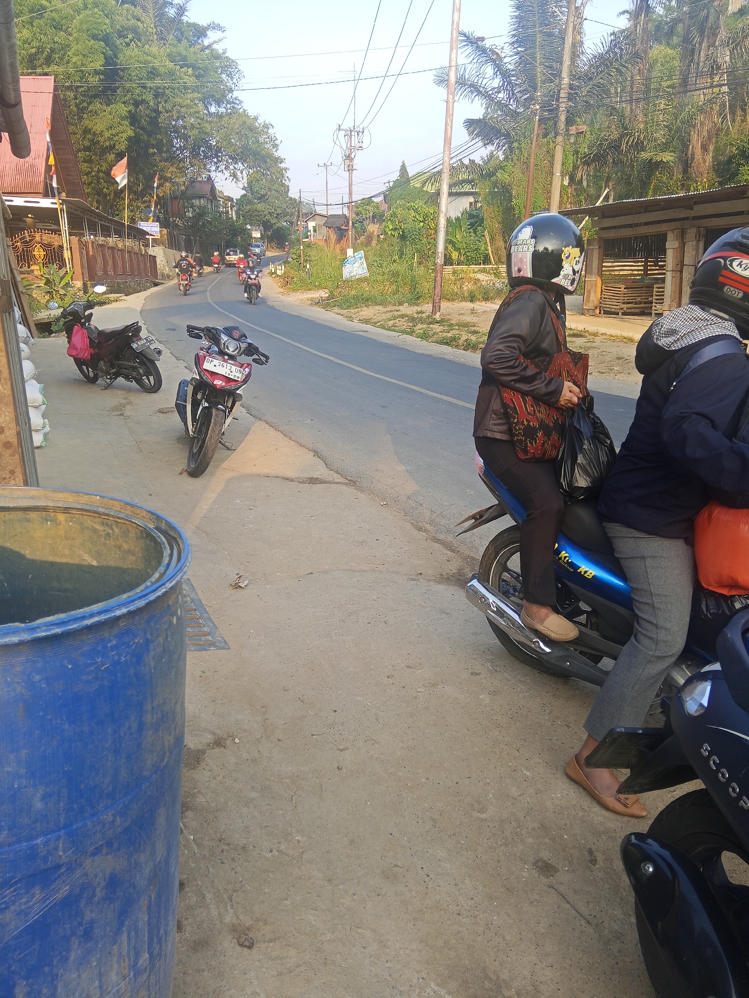 In Batu Papan, Indonesia, an outdoor scene unfolds on a sunlit day, depicting a paved road lined with vegetation and buildings. A blue plastic barrel stands prominently in the foreground. Several motorcycles are parked or in motion. One motorcycle, red and white, is parked on the side of the road. Two individuals are seated on another motorcycle, the passenger wearing a helmet with cartoon decals, a dark jacket, and carrying a patterned bag. The rider is partially visible in profile. Further down the road, other motorcycles and a white car are visible, suggesting a typical day in the area. The environment is characterized by a mix of natural elements like trees and man-made structures, creating a rural or semi-urban atmosphere.