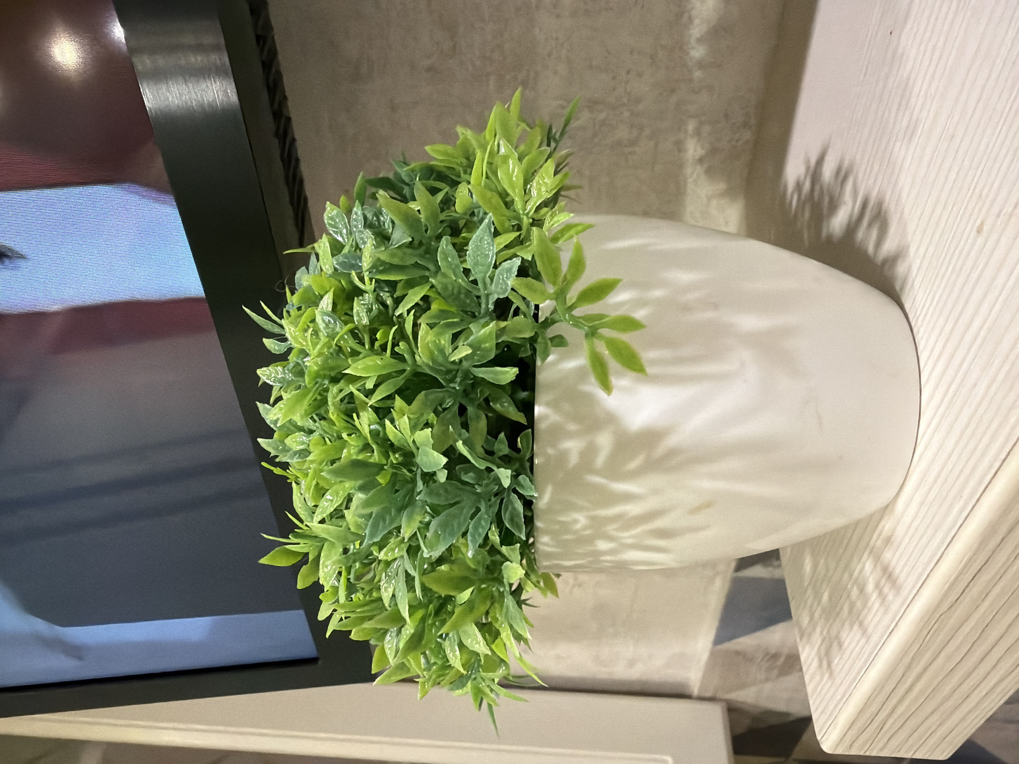 The media file shows a close-up, rotated view of a white ceramic vase filled with artificial green foliage. The vase has a smooth, rounded shape and is positioned on a textured surface, possibly a shelf or table. To the left of the vase, a dark object with a reflective surface is visible, likely a screen or monitor. The background consists of a textured wall. Sunlight casts dappled shadows from the plant onto the vase and the wall, suggesting it is daytime. There are no people or discernible activity taking place. The scene is indoors, with no clear indicators of the specific location beyond the implied setting of a room with decorative elements. The lighting suggests the presence of natural light. There is no visible text in the media file.