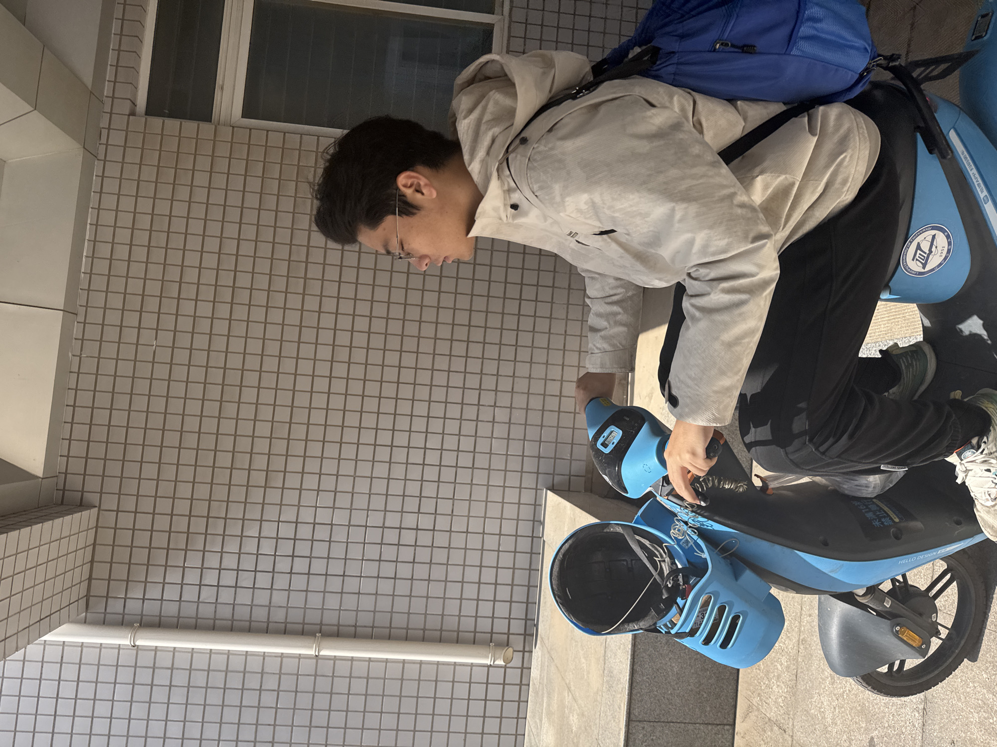 A young man is riding a blue electric scooter in an urban area, likely in Zhongtun, China. He wears glasses, a hooded jacket, and black pants, with a blue backpack on his back. The scooter has a helmet in the front basket and markings in Chinese characters, including "HELLO DESIGNE" and "1951." The background features tiled walls and a window, suggesting a building exterior.