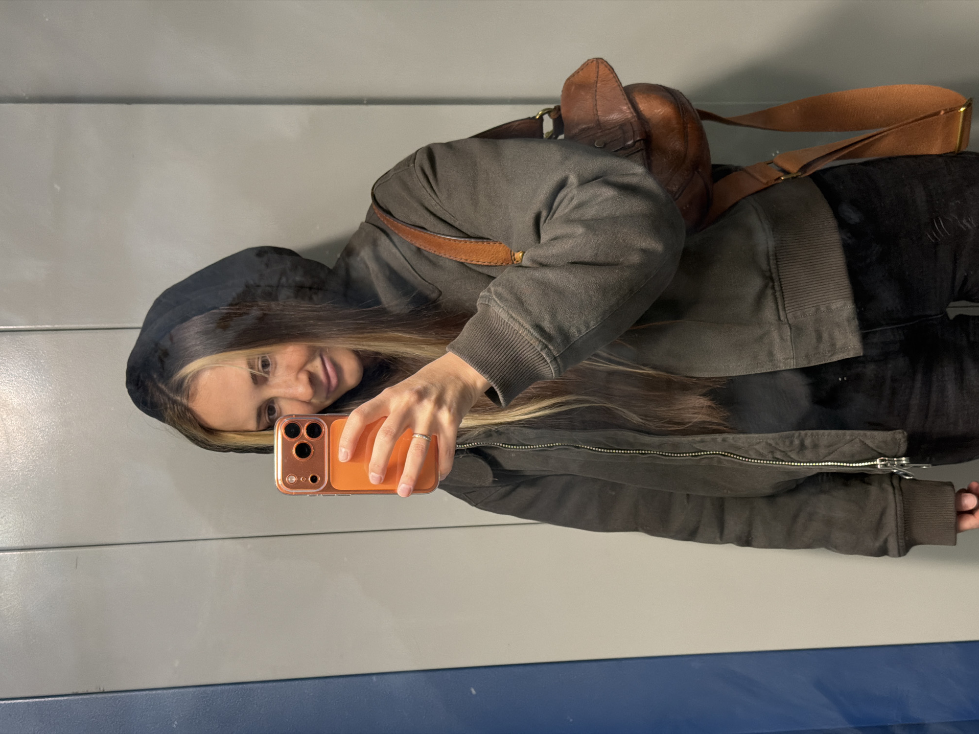 A woman is captured taking a mirror selfie, positioned slightly to the left of the frame. She has long, dark brown hair with blonde highlights and a faint smile. She is wearing a dark grey or olive green hooded jacket with a zipper, and a brown leather backpack or bag is visible over her shoulder. Her right hand, adorned with a silver-toned ring on her ring finger, holds an orange-cased smartphone to capture the reflection.
The background, as reflected in the mirror, consists of a plain light gray wall with a horizontal blue strip running along the bottom edge of the visible area. The setting appears to be an indoor public space, possibly a corridor or waiting area. No visible text is present in the image. The time of day and weather are not discernible. The location context for this image is Khimki, Russia.