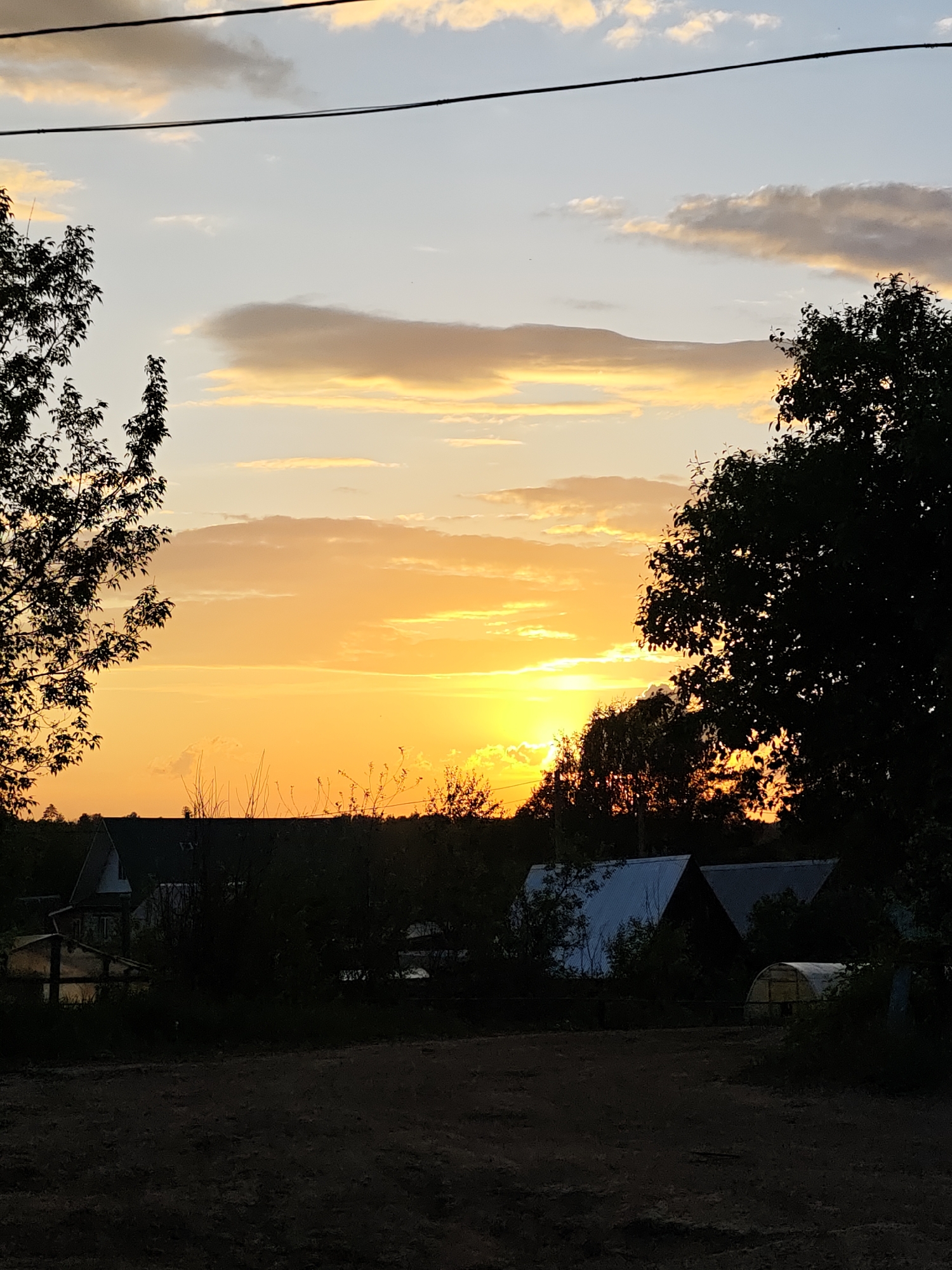 The image depicts a sunset view in the area of Уральское, Russia. The sky is a mix of light blue, yellow, and orange hues with a few clouds scattered. Trees are silhouetted on both sides of the frame. The foreground shows a dirt path with dark shrubs and what appear to be houses or structures with light-colored roofs. A power line is visible across the top of the frame.