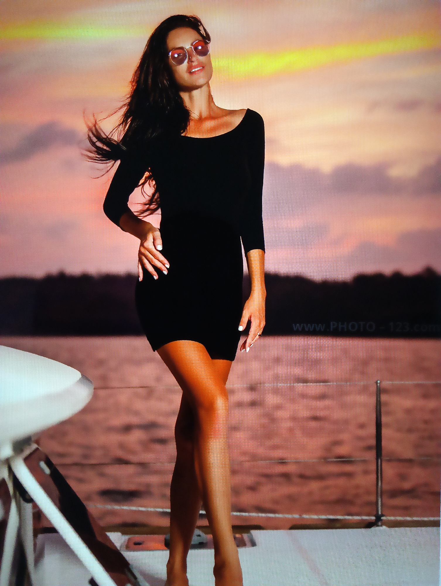 A woman stands on a boat, likely a yacht, with a body of water and trees in the background under a sunset sky. The woman is wearing a black dress and round sunglasses. A table with a white top and metal legs is visible on the left. The website "www.PHOTO-123.com" is partially visible on the right side of the image above the water line. The scene is suggestive of Nha Trang, Vietnam, with the background scenery reflecting a coastal or island setting common to the Nha Trang Ward.