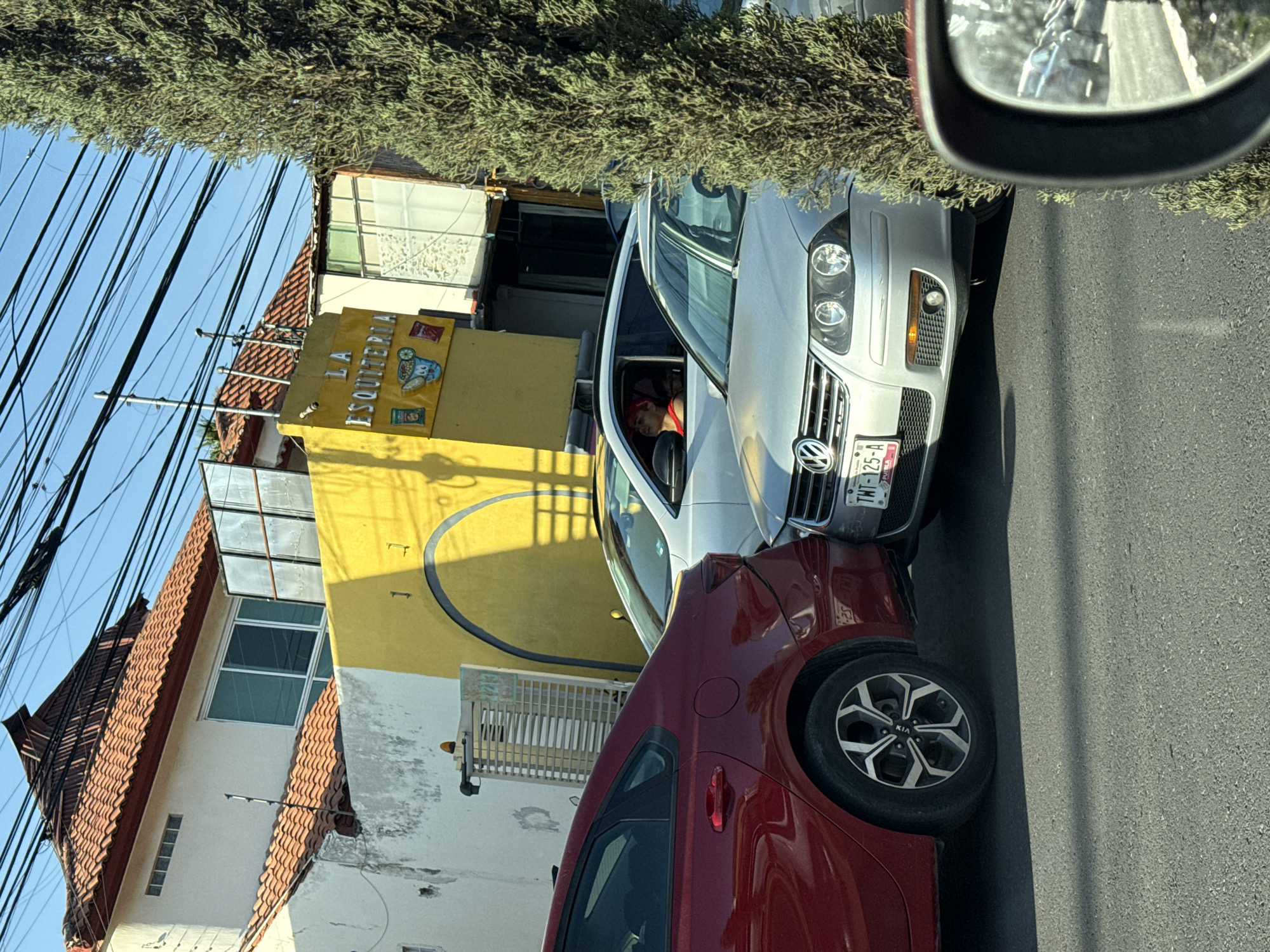 The image shows a street scene in Puebla, Mexico. A silver Volkswagen car with license plate "TNT-125-A" is parked next to a red Kia car. A person with red hair is visible in the driver's seat of the Volkswagen. Behind the cars is a building with a yellow facade. A sign on the building reads "LA ESQUITERIA." Above the yellow building is a building with a red tiled roof. Electrical wires are visible in the sky. A side mirror is present in the upper right corner of the image. The street is paved.