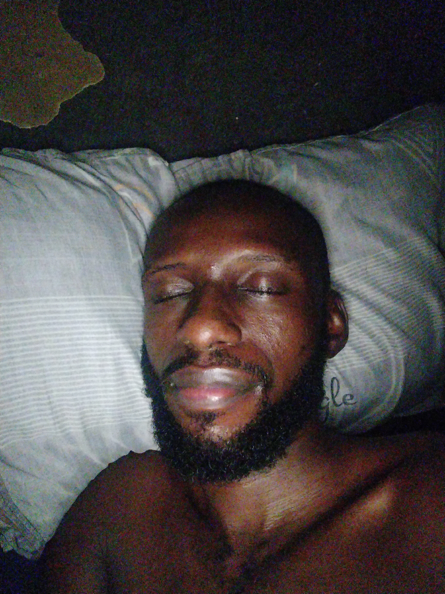 A dark-skinned, bald man with a beard is lying on a pillow with his eyes closed. The location is presumed to be indoors, based on the pillow and surrounding dark environment, but the specific location within Uyo, Nigeria cannot be determined from the image alone. The man appears to be resting or sleeping.