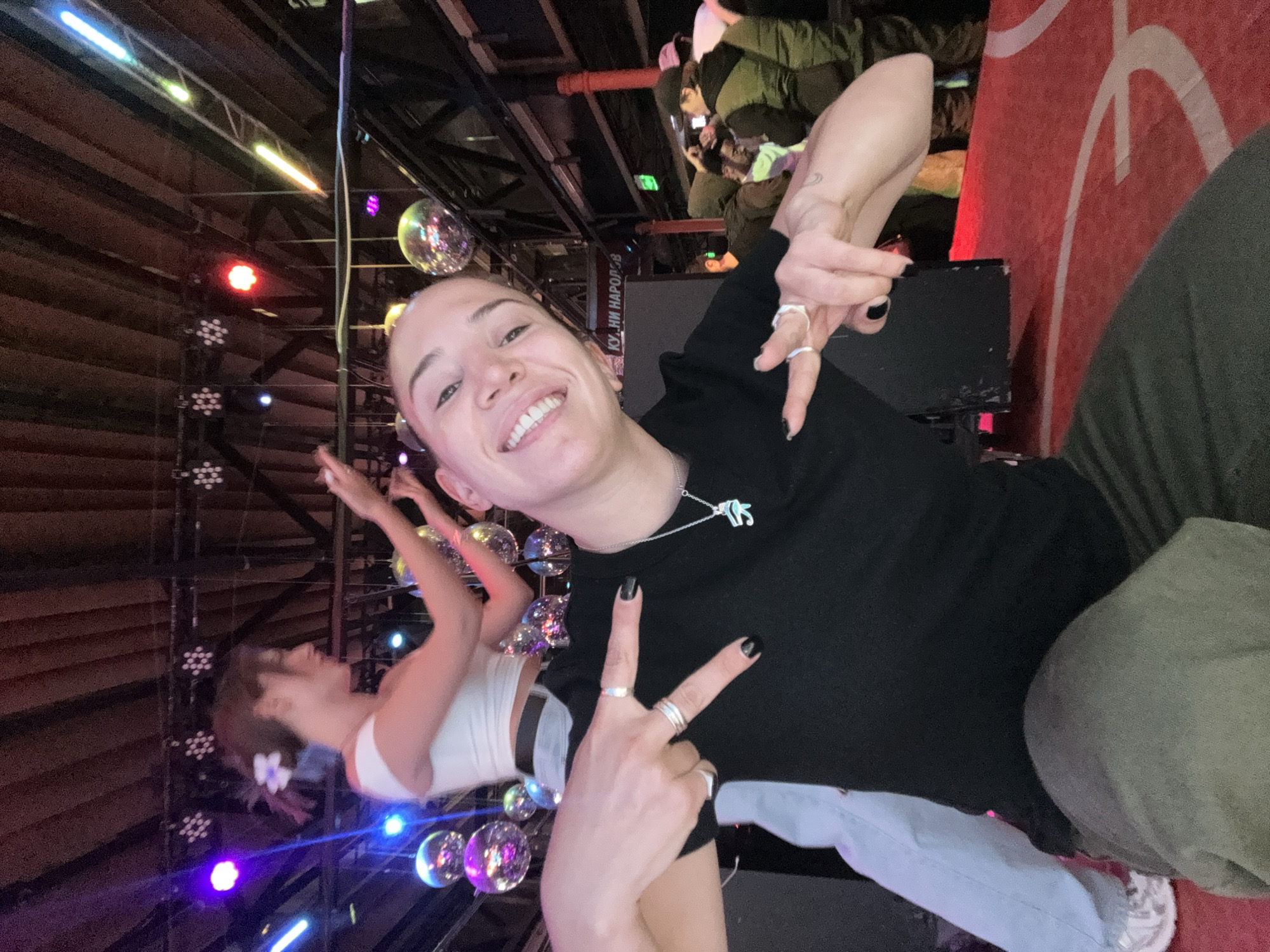 The image shows a person posing indoors, likely in a nightclub or event space in Moscow, Russia. The person is smiling and holding up two fingers in a peace sign. They are wearing a black shirt, jeans, and a necklace. There are disco balls and stage lights in the background, and another person is visible in the background performing aerial silks. The Cyrillic text visible in the background suggests the location is in Russia.