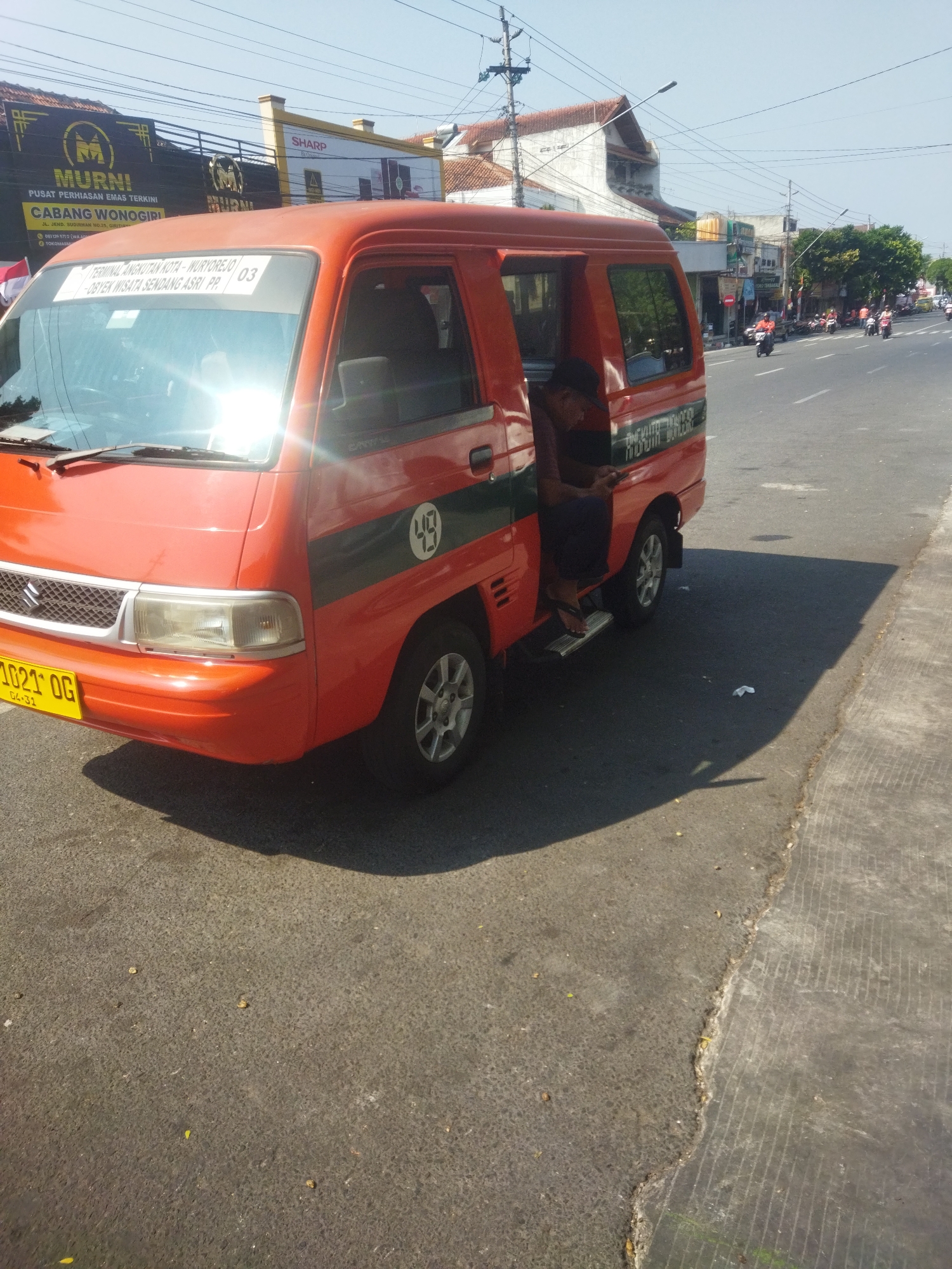 An orange passenger van with a green stripe and a white circle with a number 49 is parked on the side of a road. The van has the text "TERMINAL ANGKUTAN KOTA - WURYOREJO" and "OBYEK WISATA SENDANG ASRI PR. PP." printed on the windshield. A person is sitting in the open doorway of the van, facing away from the camera. The van has a yellow license plate that reads "1021 OG." In the background, there are buildings, trees, and power lines, with a few motorcycles visible on the road. The scene appears to be in a city. The presence of "WONOGIRI" on a sign in the background suggests the location is Wonogiri, Indonesia.