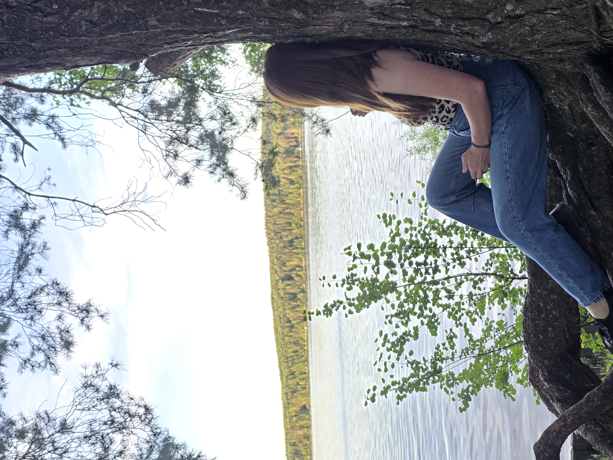 A young woman with long brown hair sits with her back against a large tree trunk on the shore of the Kama River. She is wearing a leopard print tank top and blue jeans. Her legs are crossed, and she is looking out at the water and the forested shore across the river. The water is calm, and the sky is bright with a few clouds. The tree has exposed roots that spread out along the ground. The overall scene is peaceful and serene, with the natural beauty of the landscape being a prominent feature.