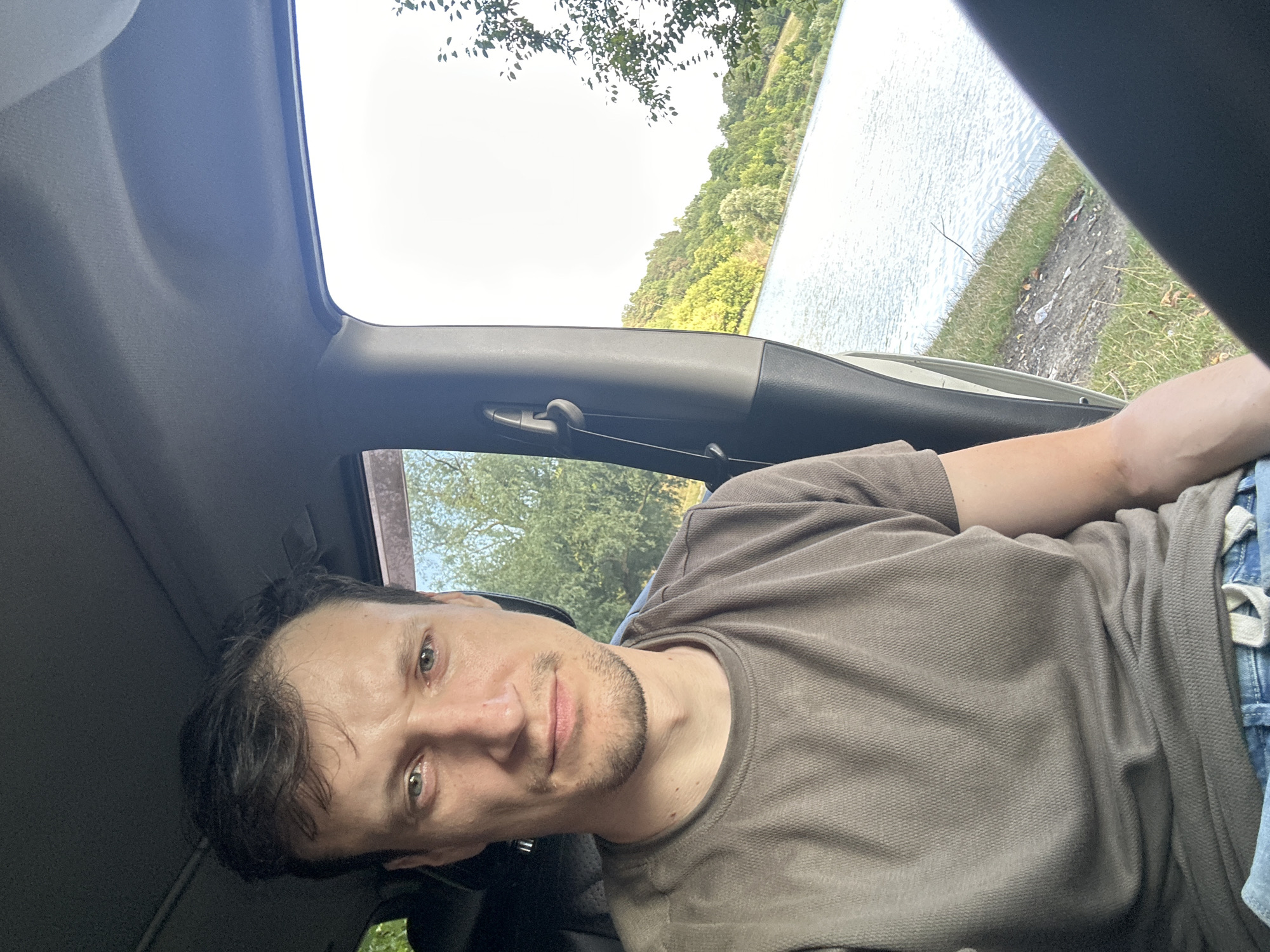 A man sits in the driver's seat of a car, looking directly at the camera. He has short, dark hair and is wearing a grey t-shirt and blue jeans. The car's interior is visible, including the dashboard and headliner. Outside the car, a body of water is visible, bordered by a grassy shoreline and a tree-covered hill. The location is identified as Makiv, Ukraine.