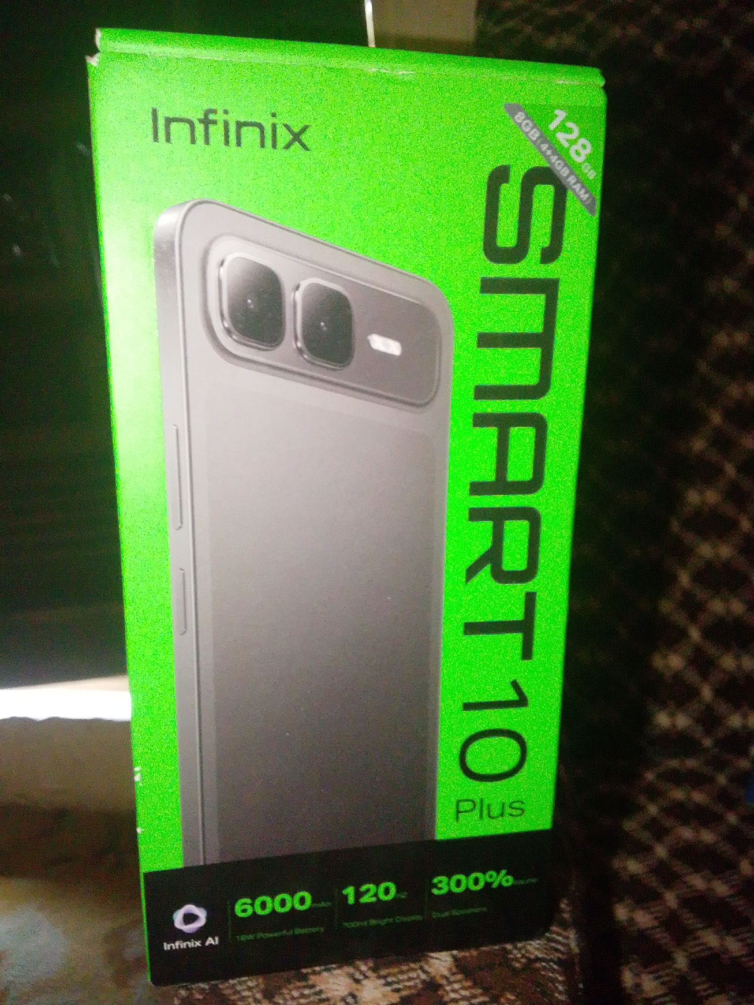 The image shows a close-up of a green Infinix Smart 10 Plus mobile phone box, which is partially obscuring the phone itself. The box is oriented vertically and angled towards the viewer. Text on the box reads "Infinix", "SMART 10 Plus", "128GB (8GB+4GB RAM)", and smaller text at the bottom details features like "6000 mAh", "120 Hz", "300%", and "Infinix AI". The phone is silver and appears to have a dual-camera system. The background is dark with a patterned fabric. This scene is located in Jalingo, Nigeria.