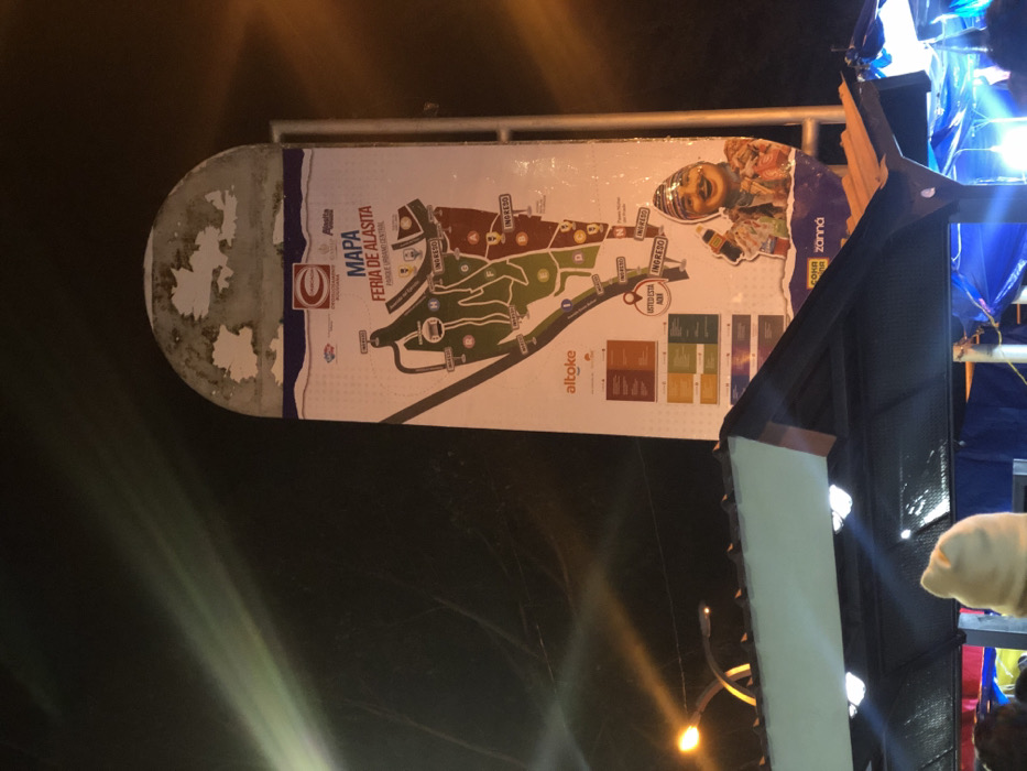 A somewhat deteriorated map displays the Feria de Alasita in Parque Urbano Central. The map highlights the layout of the fairgrounds with labeled sections and marked entrances ("INGRESO"). A corner of the map features an advertisement or promotional image with the text "Co zonná". The sign is mounted on a metal frame and positioned outdoors, with parts of adjacent structures visible to the right. "Altoke" is printed on the sign. The sign is tilted.