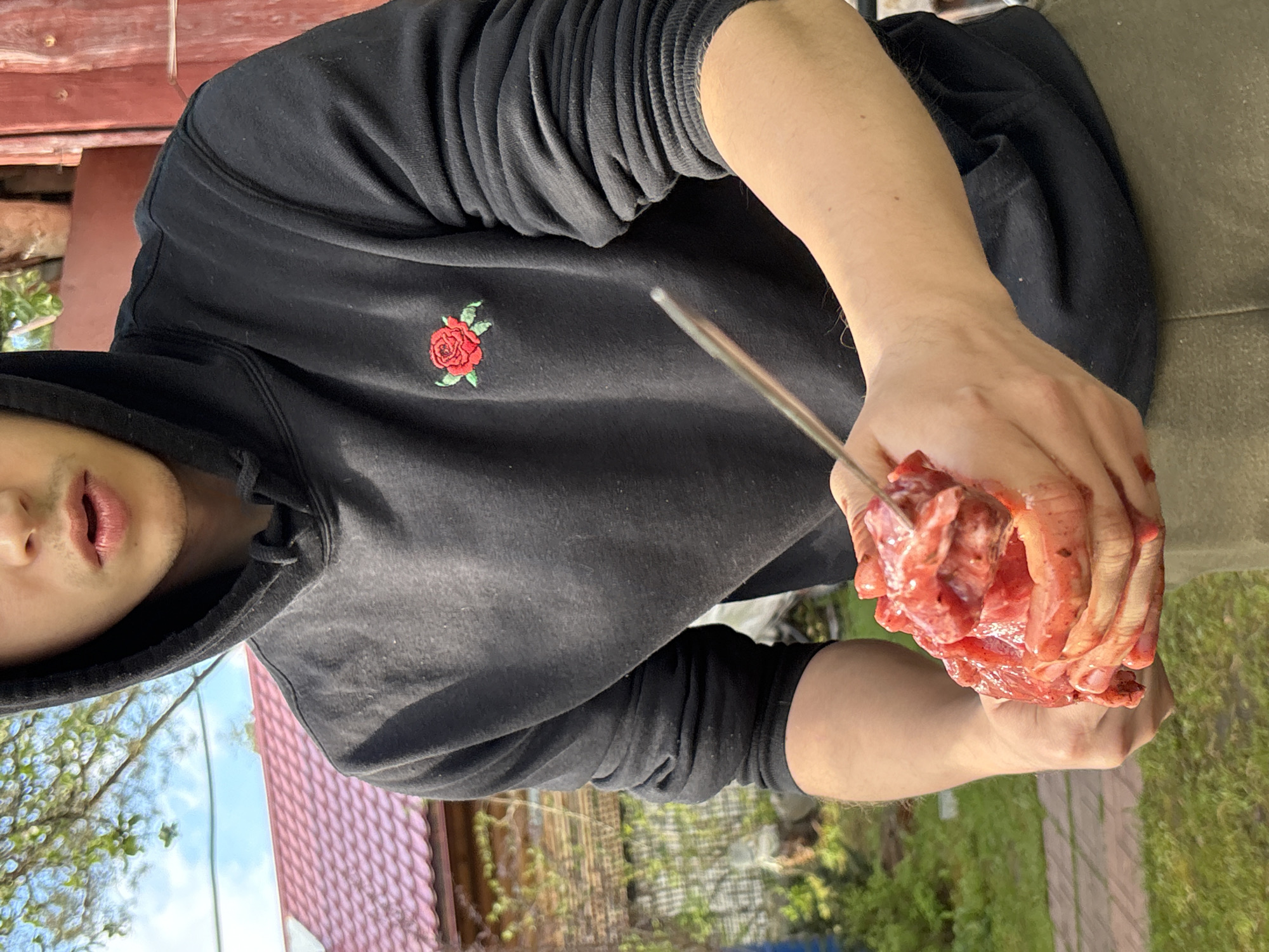 A person wearing a black hooded sweatshirt with a red rose emblem is holding a chunk of raw meat impaled on a metal skewer. The background shows a building with a red roof, trees, and grass. The location is potentially Ленинградская, Russia, although this cannot be definitively confirmed from the image. The person is not visibly interacting with anyone else.