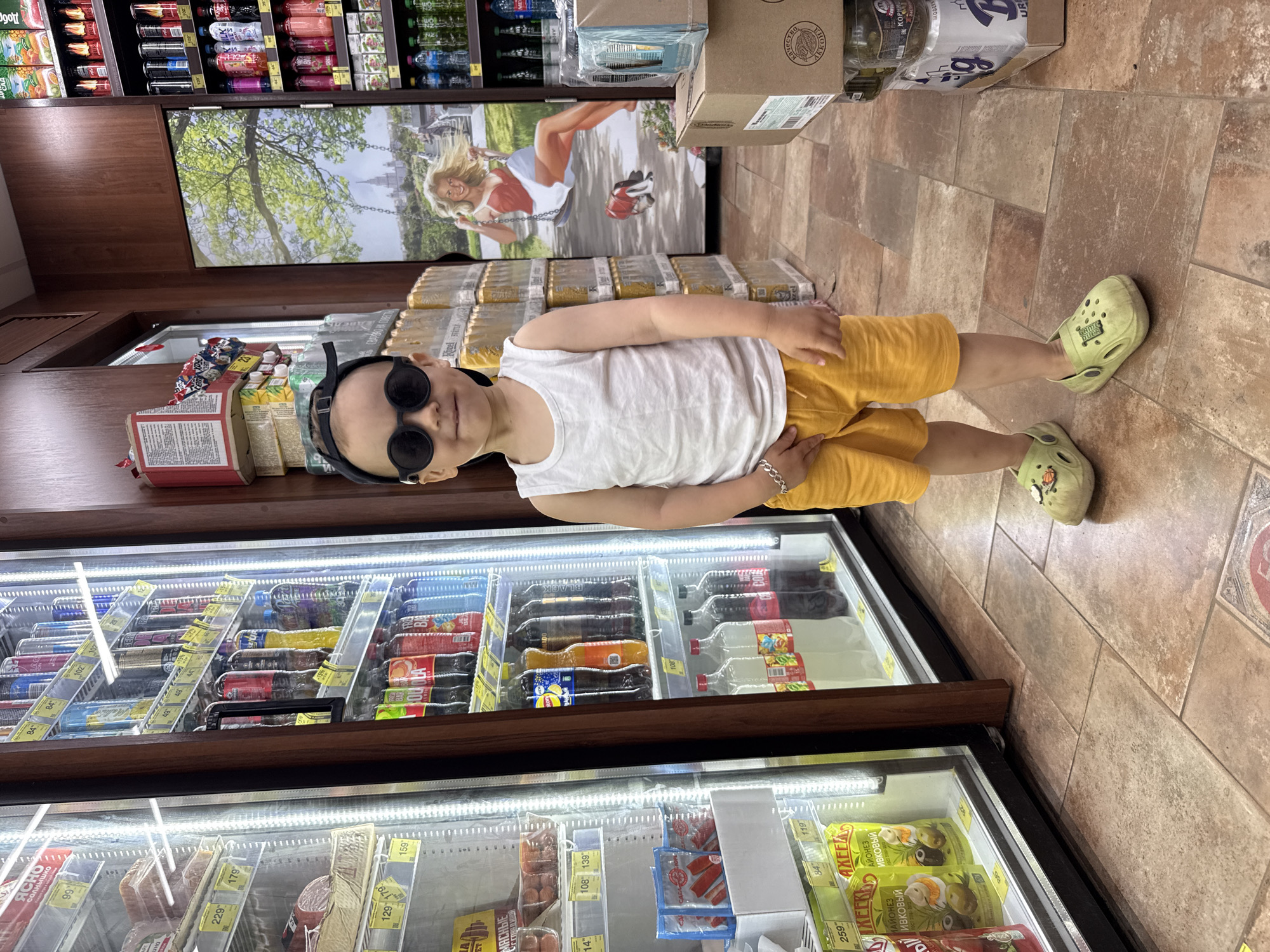 In the city of Rusanovo, Russia, a young boy stands in the aisle of a convenience store. He is wearing a white sleeveless shirt, yellow shorts, sunglasses, and light green Crocs. He is facing the camera with a neutral expression. Behind him, to his left, is a refrigerated display case filled with drinks and food items. To his right, there are shelves stocked with canned beverages and boxes of goods. A large mural depicting a woman on a swing against a backdrop of trees and a castle is visible on the wall behind him. The floor is tiled in a neutral brown color. The overall lighting suggests an indoor setting, possibly a brightly lit convenience store.