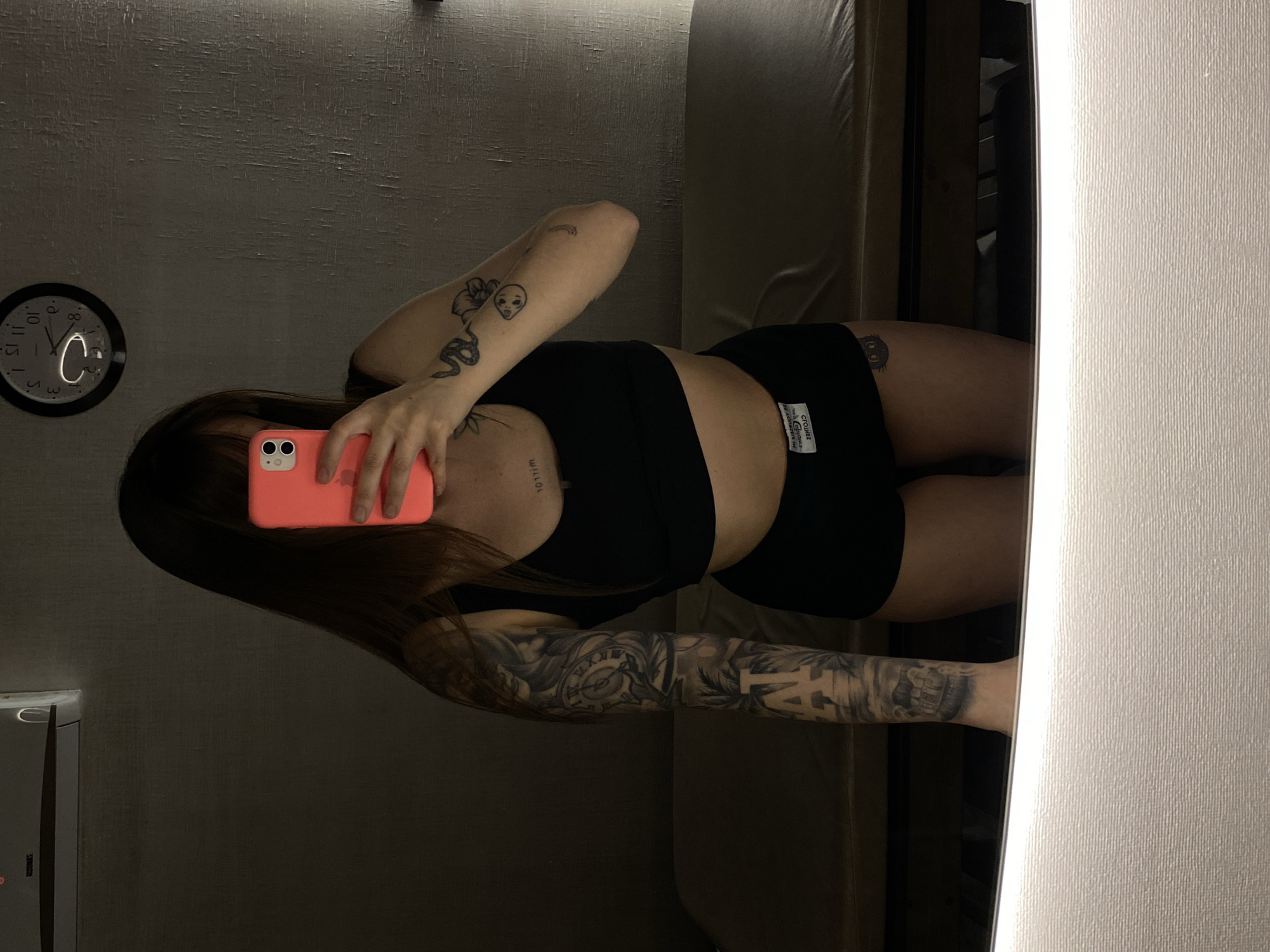 A person is seen in a mirror selfie, oriented 90 degrees clockwise from upright. They have long dark hair and are wearing a black crop top and black shorts.
Numerous tattoos are visible on their arms and body. On the right upper arm, there is a snake, a flower, a small alien-like face, and a tiny rainbow. The left arm features a full sleeve tattoo with a clock face displaying Roman numerals (I, V, X, L), foliage, and what appear to be the letters "LA." A small black cat tattoo is visible on the right hip/thigh. Faint text, possibly "willow" or similar script, is tattooed on the chest/collarbone area.
The person holds a coral/pink smartphone with dual cameras, using it to capture their reflection. In the background reflection, a black-framed wall clock with a white face shows the time to be approximately 11:55. A bright "C"-shaped reflection of a light source is on the clock face.
The setting appears to be an indoor room with textured, possibly wallpapered, walls. The lighting is dim, suggesting evening or night. On the label of the black shorts, the text "CTOUTHEZ" and other smaller, partially illegible text are visible.
The image is located in Votkinsk, Russia.