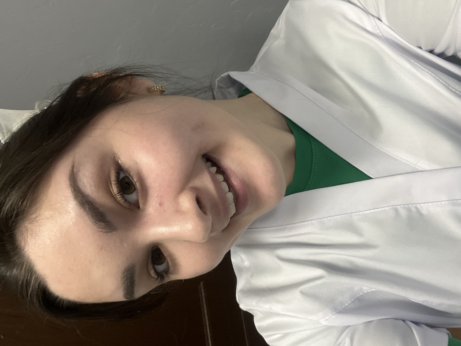 A smiling woman with brown hair and eyes is depicted from a slightly low angle. She wears a white lab coat over a green shirt. The background is a blurry mix of a wall and wooden furniture. The woman's face is well-lit, highlighting her teeth and facial features. Gold earrings are visible. Her expression is cheerful and inviting.