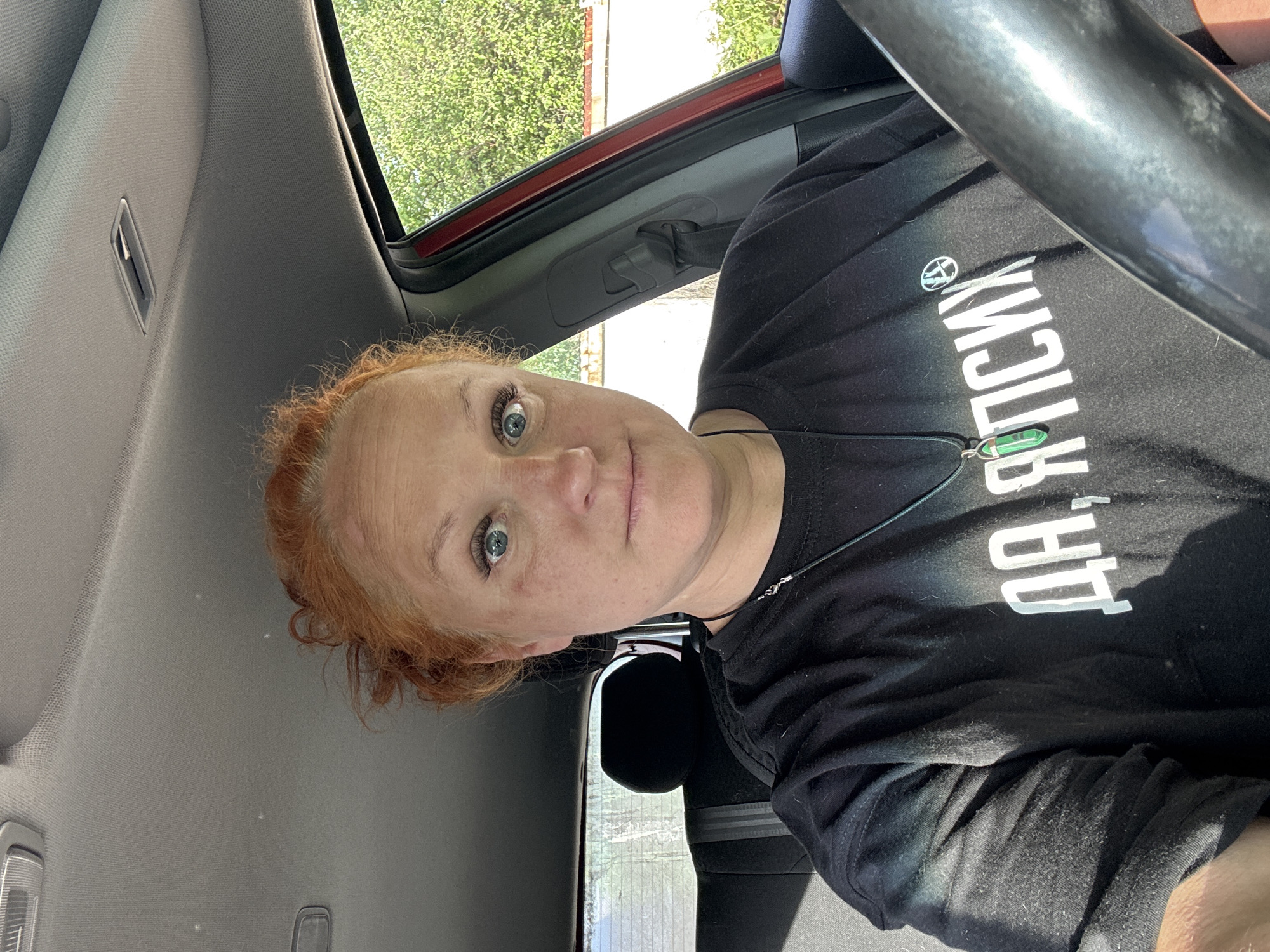 A medium shot depicts a woman inside a vehicle, likely a car, oriented towards the viewer. The woman has fair skin, red hair, and blue eyes. She is wearing a black t-shirt with white Cyrillic text "ДА, Я ПСИХ", a logo, and a necklace with a green pendant. The vehicle interior is visible, showing parts of the dashboard, the steering wheel, and the headliner. Outside the window, green foliage is visible, suggesting trees or bushes. The location is inferred to be in Chaikovsky, Russia.