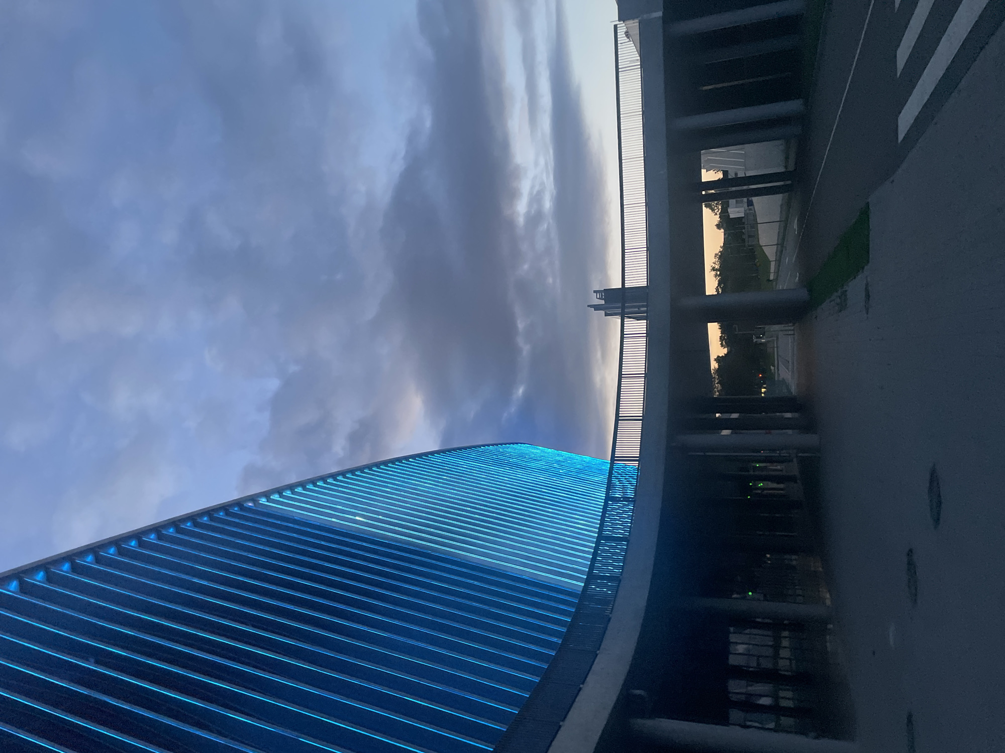 An exterior, low-angle shot captures a curved, modern building illuminated with bright blue vertical lights against a dusky sky filled with dark, dramatic clouds. A multi-level walkway or bridge with metal railings encircles the building's base, supported by large concrete columns. The walkway casts shadows on the ground below, which appears to be a paved area with visible manhole covers and a pedestrian crossing marked with white stripes. The scene depicts an architectural study of light and shadow on a contemporary structure in an urban setting at twilight.