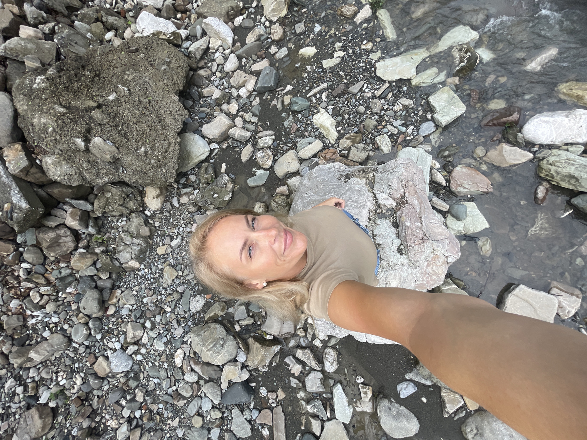 A selfie taken from a high angle shows a woman with blonde hair, fair skin, wearing a tan t-shirt, lying among rocks and water, presumably in a stream or river. The environment appears to be a natural, rocky riverbed. The location is likely in the vicinity of Sochi, Russia. The interaction is the woman taking a photograph of herself in a natural setting.