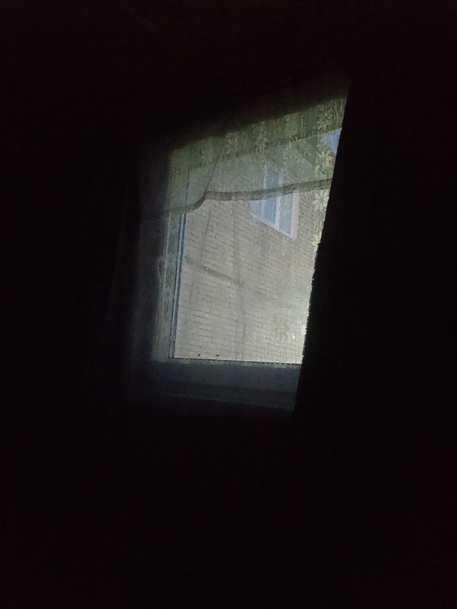 The image shows a dark, dimly lit interior looking out through a window. The window pane is somewhat dirty or textured, obscuring a clear view of the outside. Through the window, a brick building is visible, suggesting an urban or suburban environment. The brickwork appears light-colored and somewhat weathered. A portion of a window with a white frame is discernible on the building. The exterior also shows some greenery, possibly trees or bushes, which are blurred due to the distance and the condition of the window. On the right side of the image, a dark, textured surface, likely a curtain or fabric, partially covers the window and frames the view. The overall lighting suggests it is either nighttime or a very overcast day, with the interior being significantly darker than the exterior. The location is specified as Platoshino, Russia, but no specific details about the building or its surroundings that would confirm this are visible in the image itself.