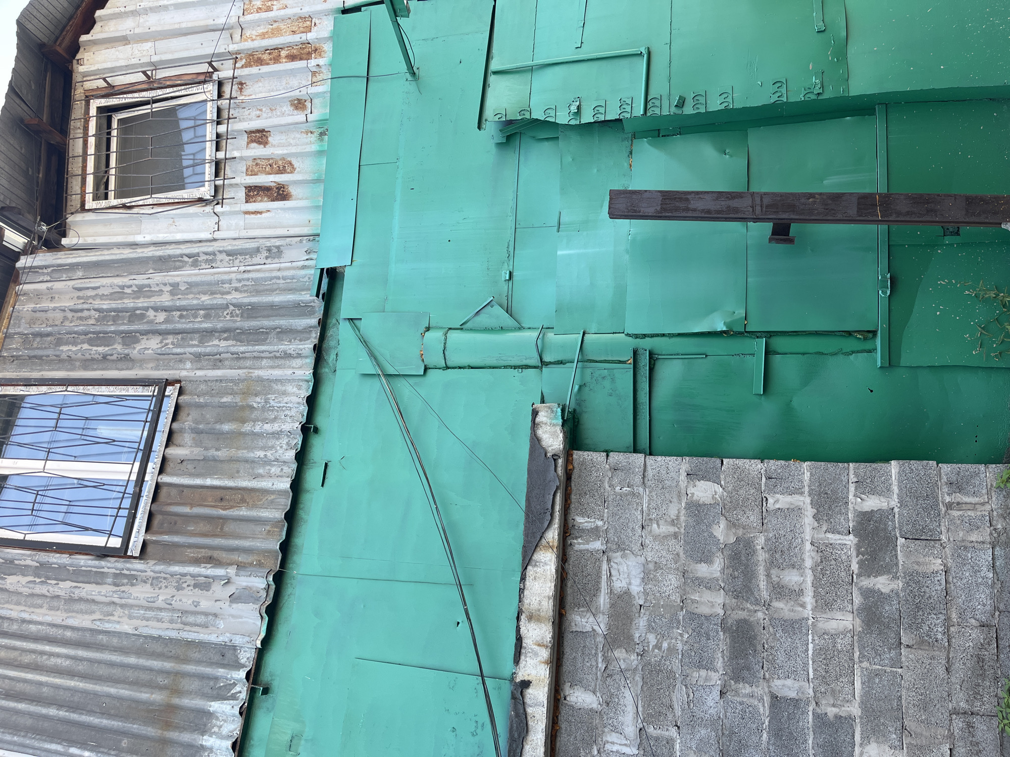 The image shows a close-up, low-angle view of the exterior of a building with a corrugated metal roof and walls, and a green metal wall on the lower section. Two windows with security bars are visible on the upper part of the building. The corrugated metal appears weathered and rusty in places. The green metal wall is constructed from overlapping panels and shows some signs of wear and tear, including dents and scratches. A section of grey concrete block wall is visible on the left side of the image, partially covered by the green metal wall. A dark, vertical post stands in front of the green metal wall, towards the right side of the image. The scene suggests an industrial or utilitarian setting, possibly an outbuilding or a less well-maintained structure in a part of Krasnoyarsk, Russia. There are no people or animals visible in the image. The overall impression is one of disrepair and age.