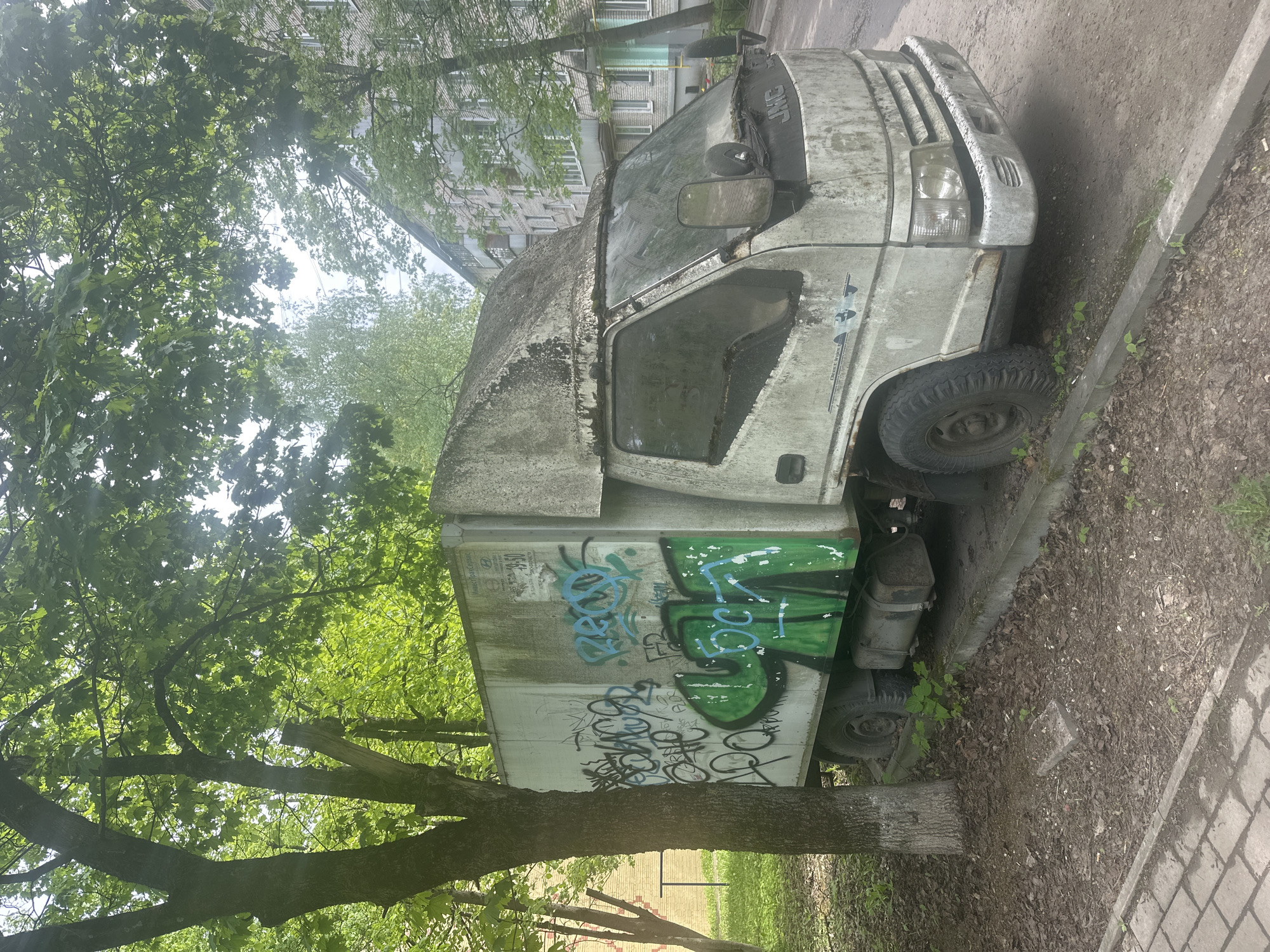 An abandoned, graffiti-covered, white commercial truck sits parked beside a tree and a concrete sidewalk in Selyatino, Russia. The truck, seemingly dilapidated, is parked along the edge of a paved area covered with loose dirt and scattered leaves. A building is partially visible in the background, obscured by the dense foliage of several trees. The overall scene suggests neglect and urban decay.
