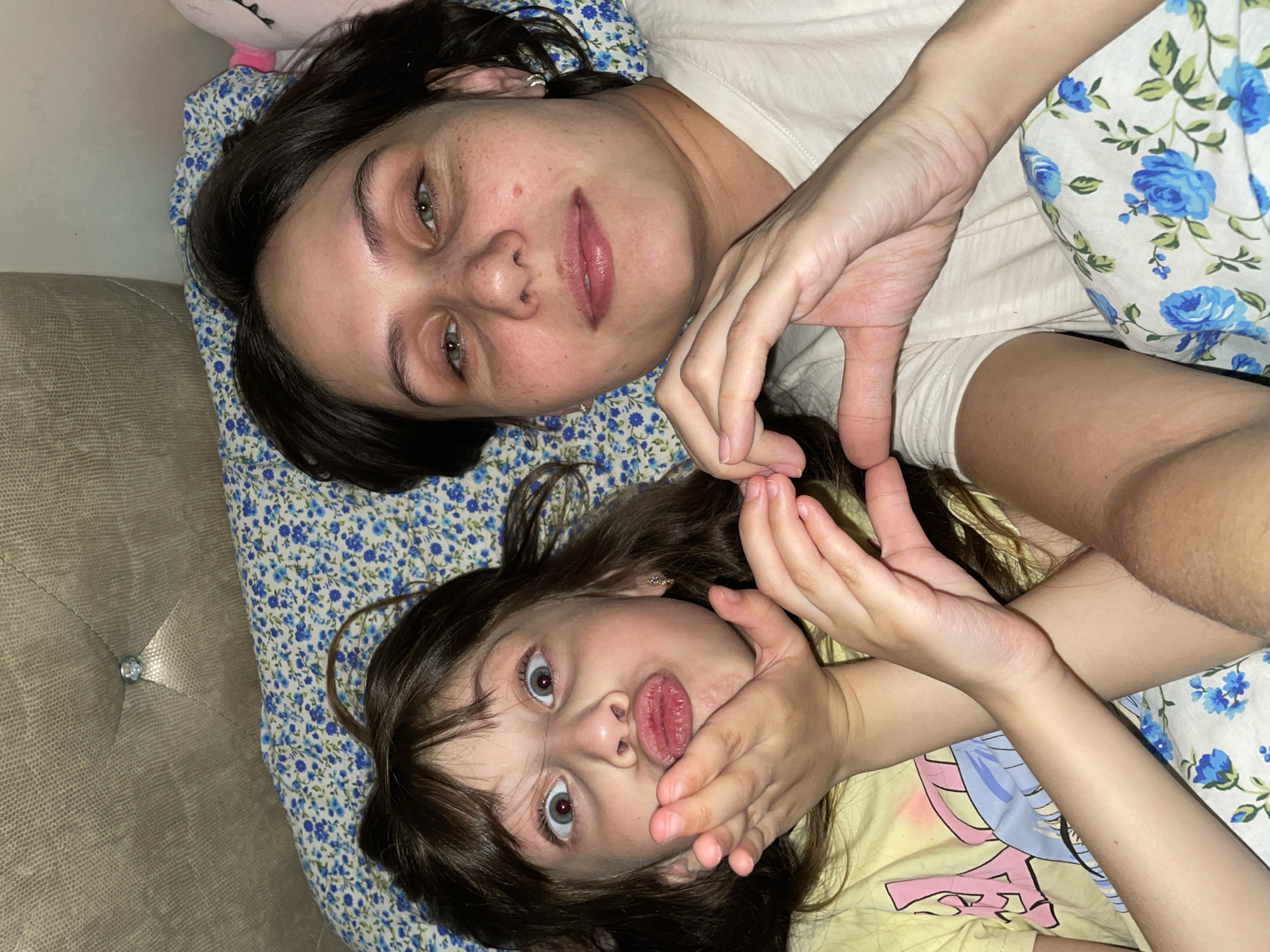 An overhead shot captures an adult woman and a child lying side-by-side, likely on a bed or sofa, engaged in a playful moment.
The adult woman, positioned towards the top of the frame, has dark hair and light-colored eyes. Her expression is calm, with a soft smile. Her face shows some natural blemishes or freckles, and she has a subtle nose piercing and earrings. She is wearing a plain, light-colored t-shirt. Her right hand is visible, curved to form one half of a heart shape.
Below her, the child, also with dark hair and wide, light-colored eyes, makes a humorous face with pursed lips. Her hands are held near her face, with her left hand joining the adult's to complete the heart gesture. The child is wearing a yellow t-shirt featuring a colorful graphic design, possibly an anime or cartoon character.
Both individuals are resting on bedding adorned with a blue and white floral pattern, depicting small blue flowers and green leaves. To the left of the frame, a textured, light brown or grey upholstered surface, possibly a tufted headboard or sofa, is visible, featuring a decorative button. A glimpse of a pink object, likely a plush toy, can be seen near the adult's head.
The scene portrays a warm and affectionate interaction, likely between a parent and child, highlighting a moment of joy and connection. The setting is an indoor domestic environment. No discernible text is visible in the image.