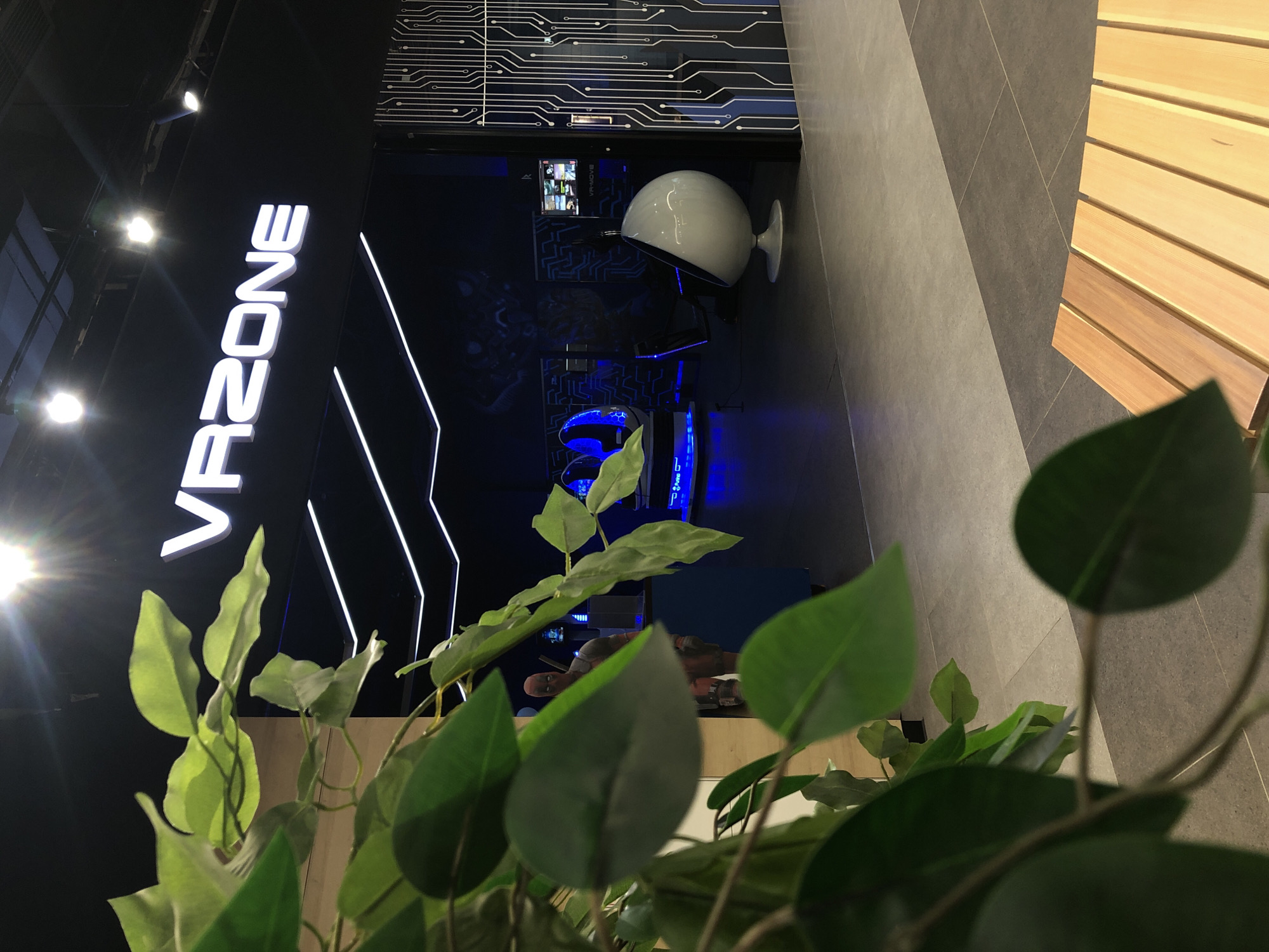 The image shows the interior of a VR zone in Izhevsk, Russia. The most prominent subject is the illuminated white text "VR ZONE" mounted vertically on a black wall. To the right of the text, abstract white lines resembling a circuit board are visible on the wall.
In the background, the VR zone features several VR setups. One notable object is a large, white, egg-shaped VR headset on a stand. Another is a darker, blue-lit VR headset, possibly for multiple users. In the middle distance, a screen displays several smaller video feeds. A figure, resembling the character Deadpool, is visible in the background, resting on a ledge.
The setting appears to be indoors, with a modern, futuristic aesthetic suggested by the circuit board motif and the sleek design of the VR equipment. The lighting is primarily artificial, with spotlights illuminating the "VR ZONE" sign and blue accents in the VR areas. The floor appears to be a combination of dark grey tiles and a lighter, possibly wooden, material on the right side of the frame. In the foreground, out-of-focus green leaves from a plant partially obscure the view of the VR zone. The overall atmosphere is dimly lit, highlighting the illuminated elements of the VR experience.