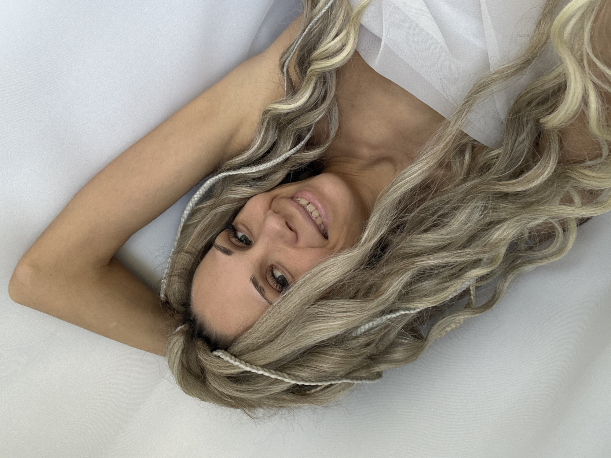 A medium-shot photograph depicts a woman lying on her back on a white surface, possibly fabric, angled from the top-left to the bottom-right. Her head is tilted towards the camera, and she is smiling, revealing her teeth. She has long, wavy blonde hair with interspersed white braids. The woman's right arm is raised and bent at the elbow. The background is an unidentifiable white material. The location is unspecified.