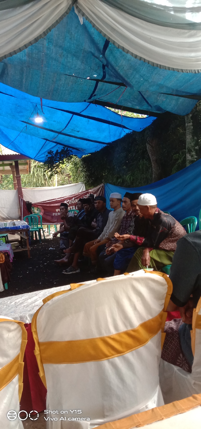 A group of men sit under a makeshift tent constructed with blue tarpaulin and white cloth. The men wear various traditional Muslim head coverings, including white caps and black hats. They are seated on a platform covered in dark material. In front of them are white chairs with gold trim. A banner and a small table with green chairs are visible in the background.