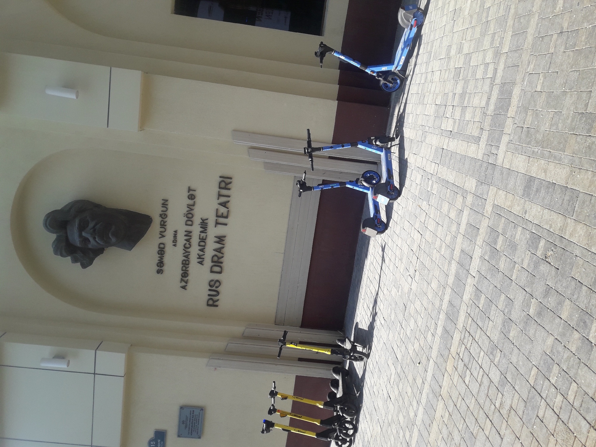 An outdoor medium shot captures the facade of a building and a row of electric scooters on a paved sidewalk. The building, painted a light yellow, features a large, dark bronze bust of a man mounted above an inscription that reads, "SƏMƏD VURĞUN ADINA AZƏRBAYCAN DÖVLƏT AKADEMIK RUS DRAM TEATRI." To the left of the bust, a sign with the number "15" is visible, and below it, a smaller plaque with text.
In front of the building, a line of electric scooters is parked. The first four scooters are yellow and black, with "Wing" branding. The remaining three scooters are blue and black, with the brand name "JET" visible on the rear wheel fender of the scooter on the far right. The scooters are parked on a sidewalk paved with light grey and beige rectangular stones arranged in a pattern. The background shows vertical wooden planks leaning against the building and a dark-colored window. Shadows from the scooters and planks are cast on the paved ground, indicating a sunny day. The image appears to have been taken from a slightly low angle.