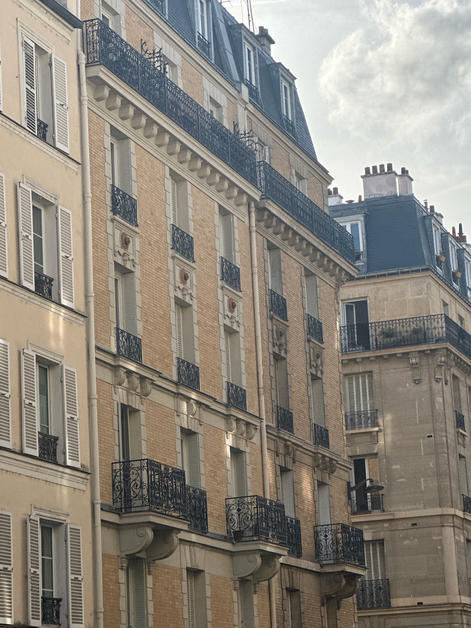 The image depicts the facades of several tall buildings in Paris, France, viewed from a low angle looking upwards. The buildings exhibit classic Parisian architecture, with tan-colored brickwork and light-colored stone accents. The buildings feature multiple stories with numerous windows, many of which are adorned with white shutters, some open and some closed. Ornate black metal railings are visible on balconies and along the edges of some of the upper floors. Chimneys and dormer windows are prominent on the dark-tiled mansard roofs. The sky above is partly cloudy with diffused sunlight. There are no people visible in the frame. The scene captures a typical streetscape in a Parisian neighborhood, focusing on the architectural details of the residential buildings.