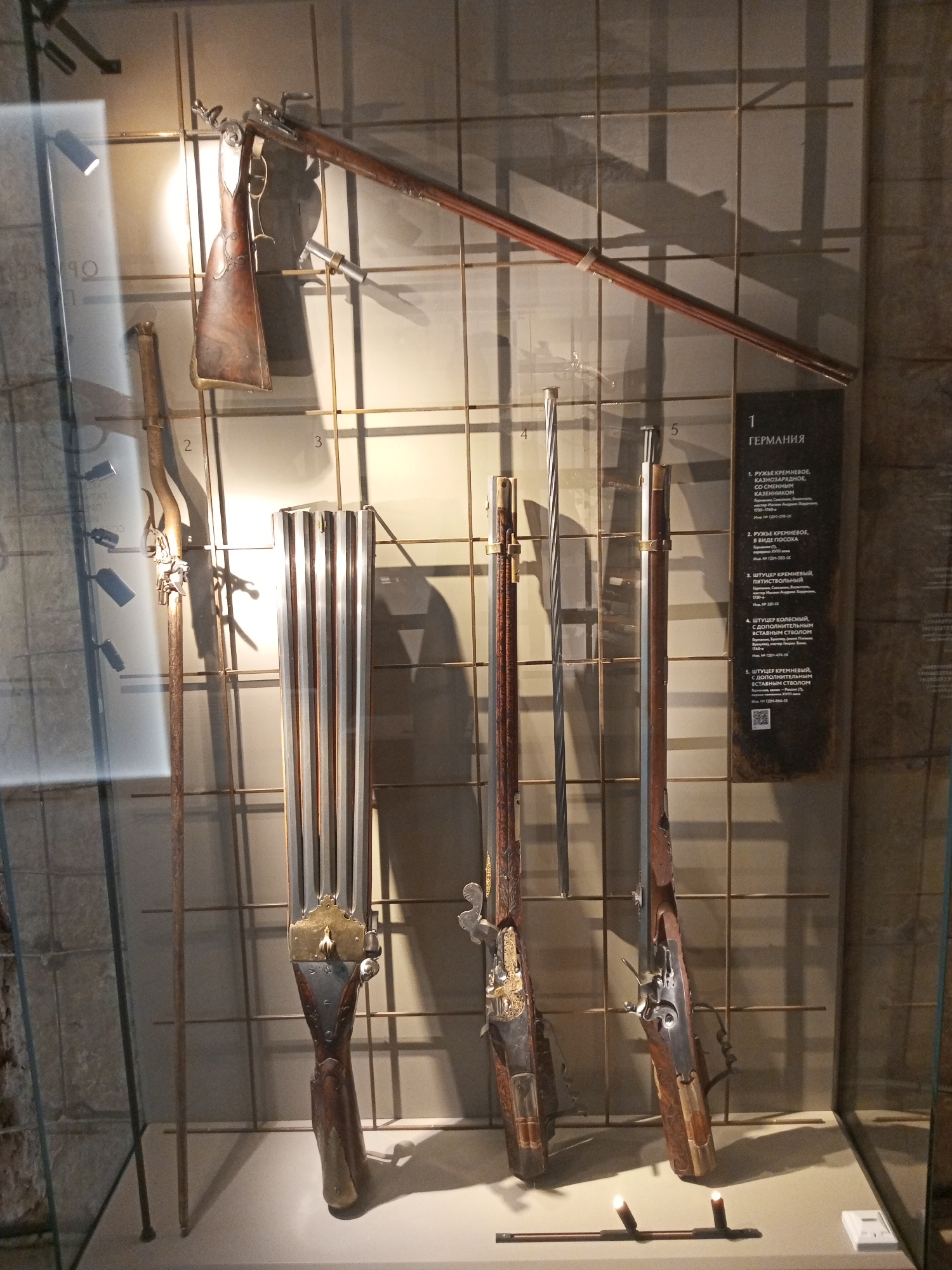 The image shows a display case filled with antique firearms, specifically muskets and rifles, labeled as German artifacts. The items are mounted on a wire grid background. From left to right, they include a long-barreled musket (possibly a fowling piece), a multi-barreled firearm (likely a volley gun or early machine gun prototype), and several muskets or rifles with ornate details. A plaque with German text next to the firearms appears to provide descriptions or historical information about them. Given the language of the placard and the style of weaponry, it is probable that this is located in a museum setting, possibly Gatchina Palace in Gatchina, Russia. The objects are under glass, and the lighting is directed from above, casting shadows.
