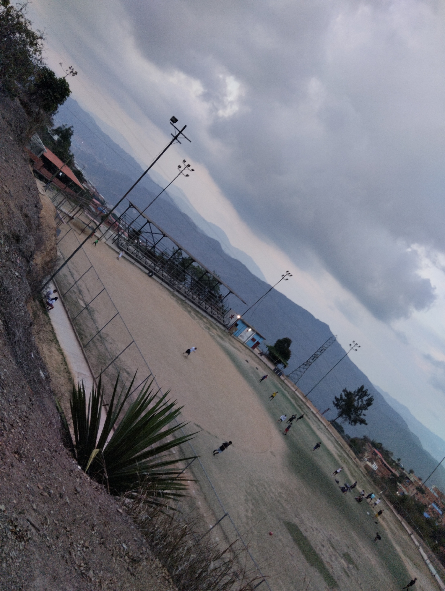 The image depicts a soccer field in Caracas, Venezuela, set against a mountainous backdrop under a cloudy sky. The field, enclosed by a chain-link fence, is occupied by several individuals engaged in what appears to be a soccer practice or game. The players are scattered across the field in various positions. Light poles are situated around the field's perimeter to illuminate it. In the foreground, a rocky hillside with vegetation provides an elevated view of the field. A few buildings are visible in the distance, blending into the urban landscape.
