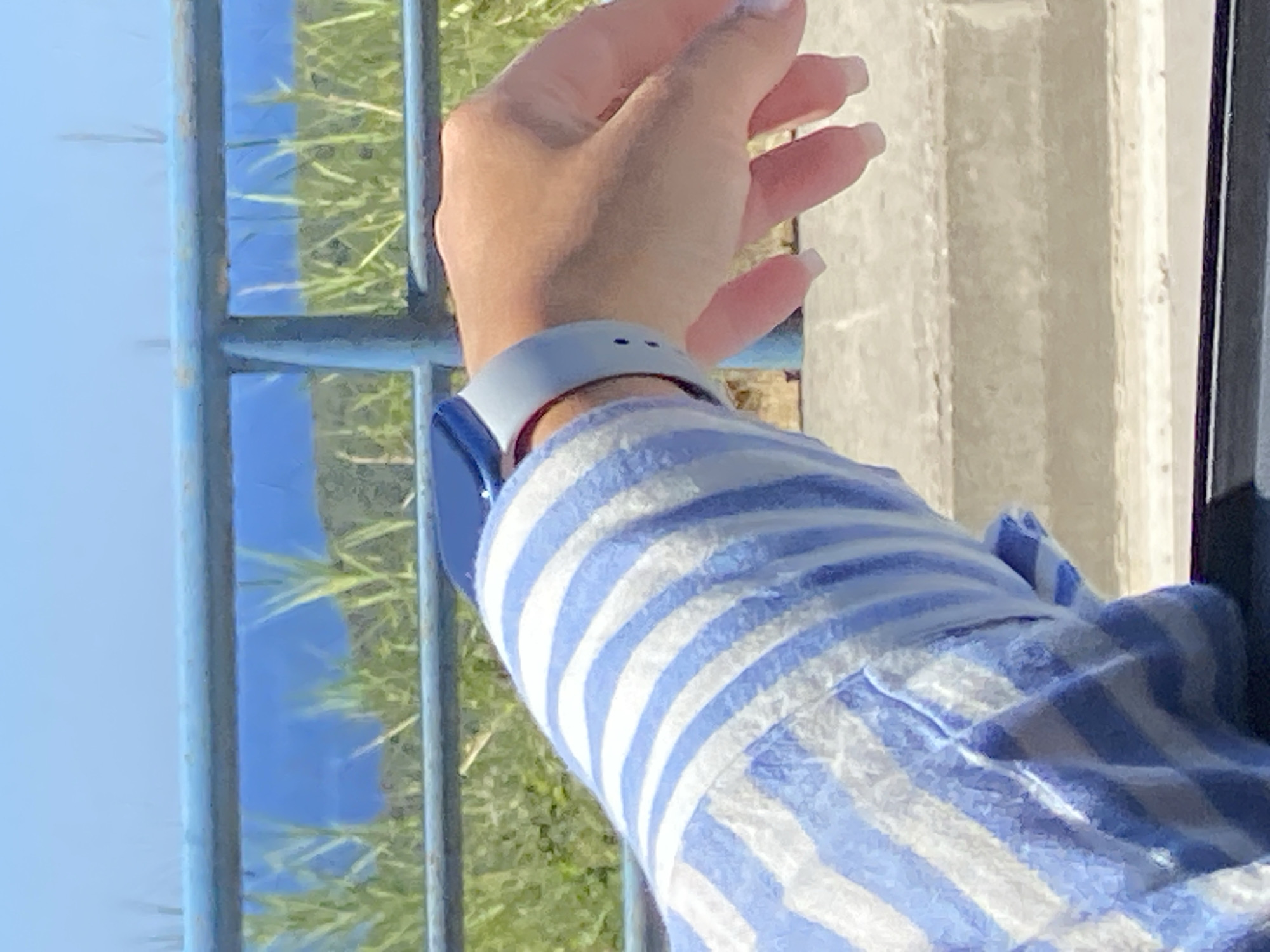 An unidentifiable person's arm, wearing a blue and white striped shirt and a light blue smartwatch, is visible from the shoulder down, extending over a blue metal railing. Behind the railing, green grass and a body of water are visible under a clear blue sky. The scene is outdoors, potentially on a balcony or overlook, with a concrete surface in the foreground. The image appears to be taken in Prijevor, Montenegro, given its proximity to the Bay of Kotor.