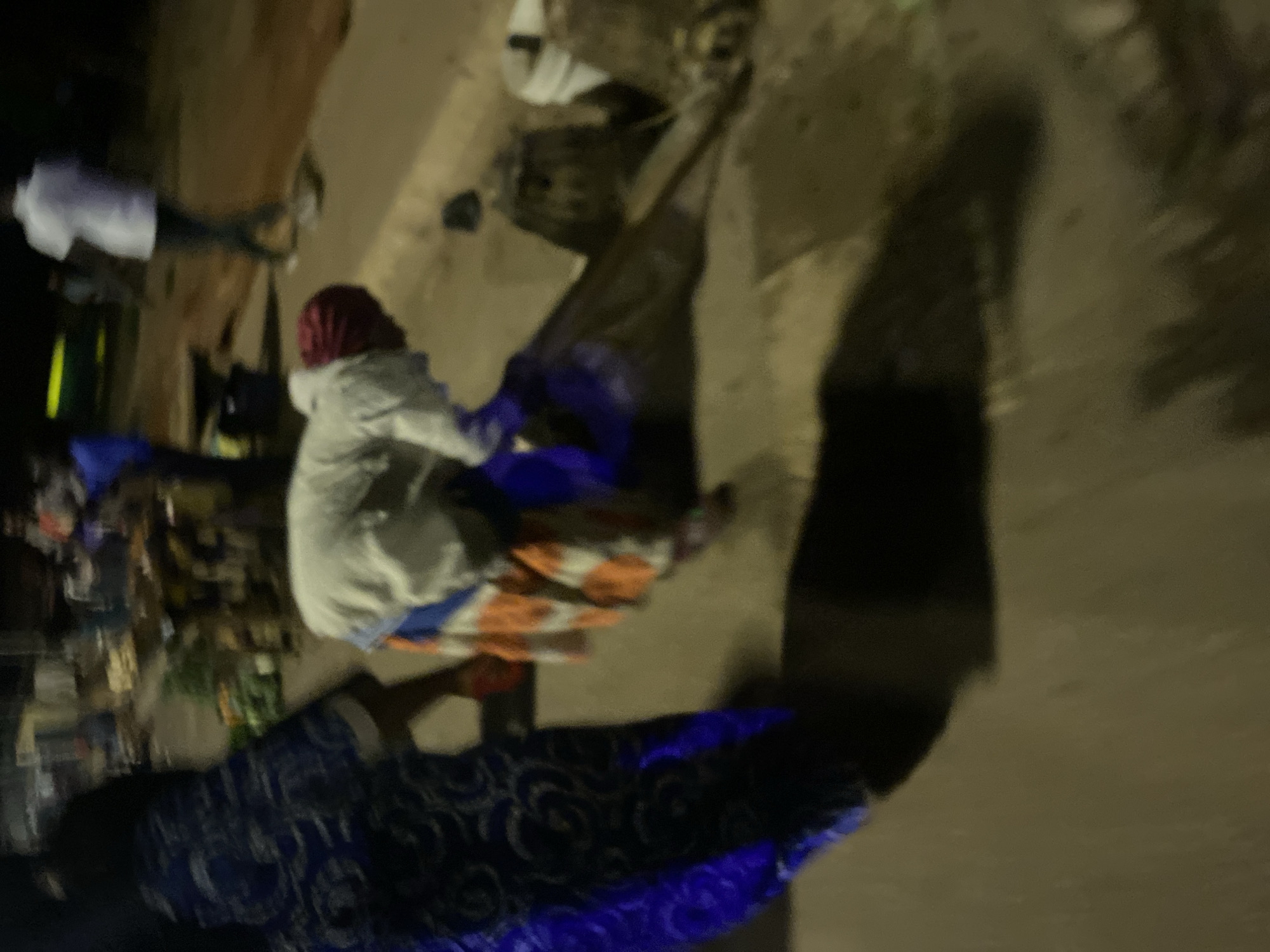 This image depicts a nighttime street scene in Bukuru, Nigeria. In the foreground, a person wearing a white jacket and an orange and white patterned skirt is bent over, appearing to pick something up from the ground. To the left, another individual is visible in a blue patterned garment, with their back to the camera. In the background, other figures are present, including one person in a white shirt and dark pants. Various objects, possibly market stalls or debris, are scattered around. The ground appears to be paved. The lighting suggests artificial streetlights are the primary source of illumination.