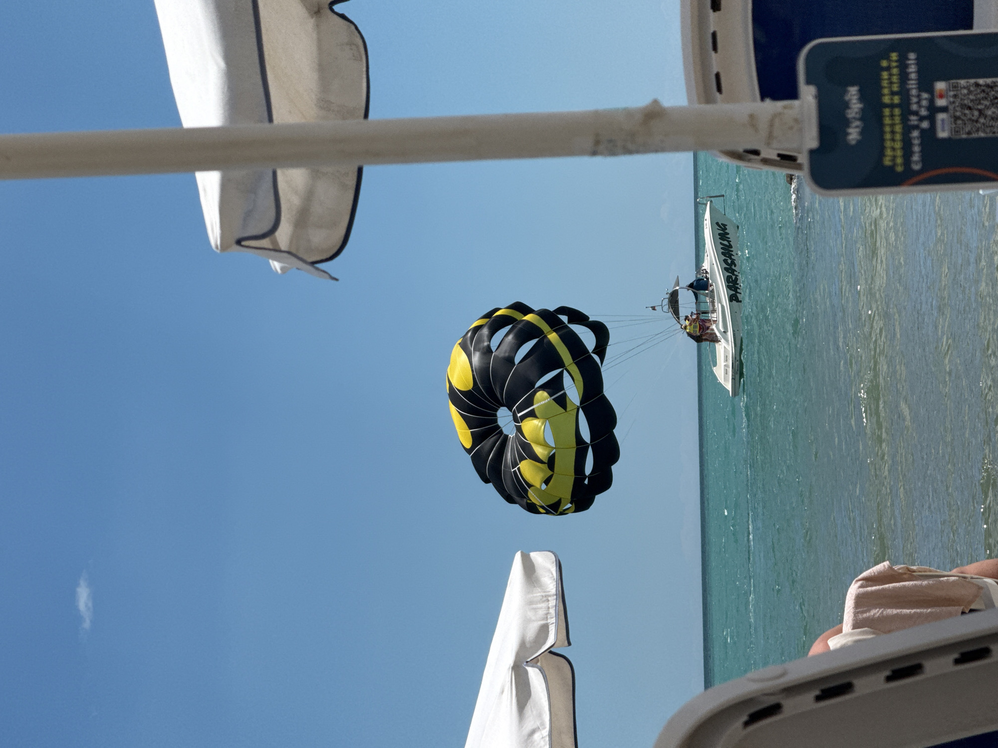 The image depicts a sunny day at a beach in Sveti Vlas, Bulgaria, with a parasailer soaring in the sky, towed by a speedboat. The parasail is a vibrant black and yellow parachute, a striking contrast against the clear blue sky. Below, the turquoise waters of the Black Sea are calm, with gentle waves lapping at the shore. A white speedboat with "PARASAILING" written on its side is visible in the distance, with people on board. Beach chairs and umbrellas are scattered along the sandy shoreline, hinting at a relaxing day by the sea. In the foreground, part of a beach chair and a white umbrella are visible, framing the scene. A sign with a QR code and text in Cyrillic and English, likely for payment or information, is also present in the foreground. The overall atmosphere is one of leisure and outdoor activity.