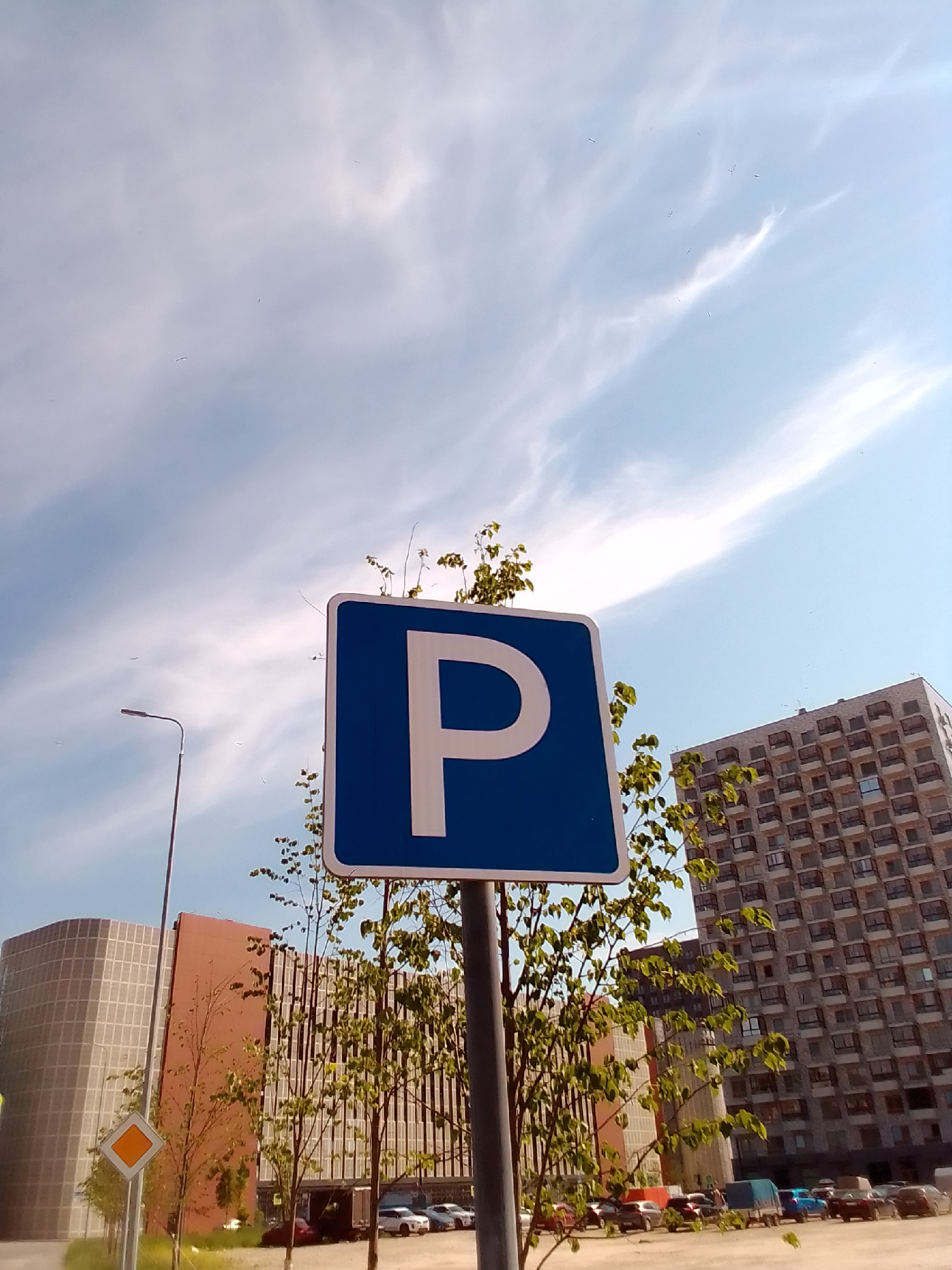 A close-up, eye-level outdoor shot captures a bright, sunny day in a city. The sky, a vibrant blue with wispy white clouds, fills the upper portion of the frame. A prominent blue parking sign with a white "P" is centered in the foreground, slightly tilted. Behind the sign, several trees with green leaves are visible, with branches reaching upwards. In the background, two modern, multi-story buildings stand tall. The building on the left is curved and covered in a grid-like facade, while the building on the right is rectangular with numerous balconies. A street light is positioned to the left of the parking sign. Below the buildings, a parking lot with several cars is apparent. A yellow diamond-shaped sign is visible to the left of the parking lot. The overall scene suggests an urban environment, possibly a residential or commercial area.