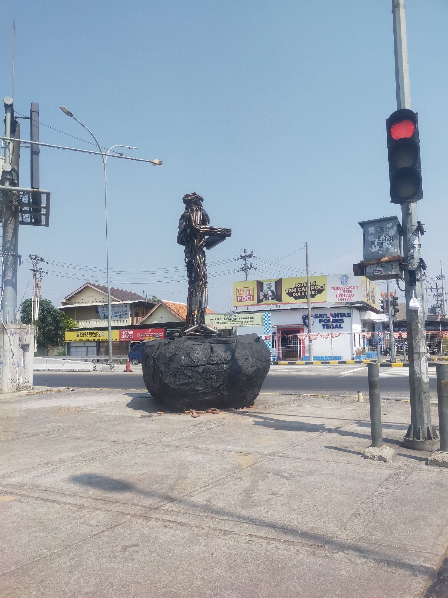 The image depicts a street scene in Lemahduwur, Indonesia, on a clear, sunny day. In the foreground, a concrete sidewalk is visible, casting long shadows from the objects and structures around it. Dominating the center of the frame is a large, dark, bulbous sculpture, possibly representing a rock or earth element, from which a metallic sculpture of a person emerges. This humanoid figure appears to be made of scrap metal, with a head, torso, arms holding an object, and legs.

To the left of the sculpture, a tall utility pole with multiple wires and what looks like a traffic signal control box is present. Further to the left, a street lamp extends over the sidewalk. In the background, several buildings line the street. One building has a banner that reads "POS TERPADU POLISI LALU LINTAS". Another building displays advertisements for "TEH POC", "BUDAYAKAN", and "SATLANTAS POLRES TEGAL". A red traffic light is illuminated on a pole to the right of the frame. The environment appears to be an urban setting with active traffic infrastructure. There are no visible people in the immediate vicinity of the sculpture.
